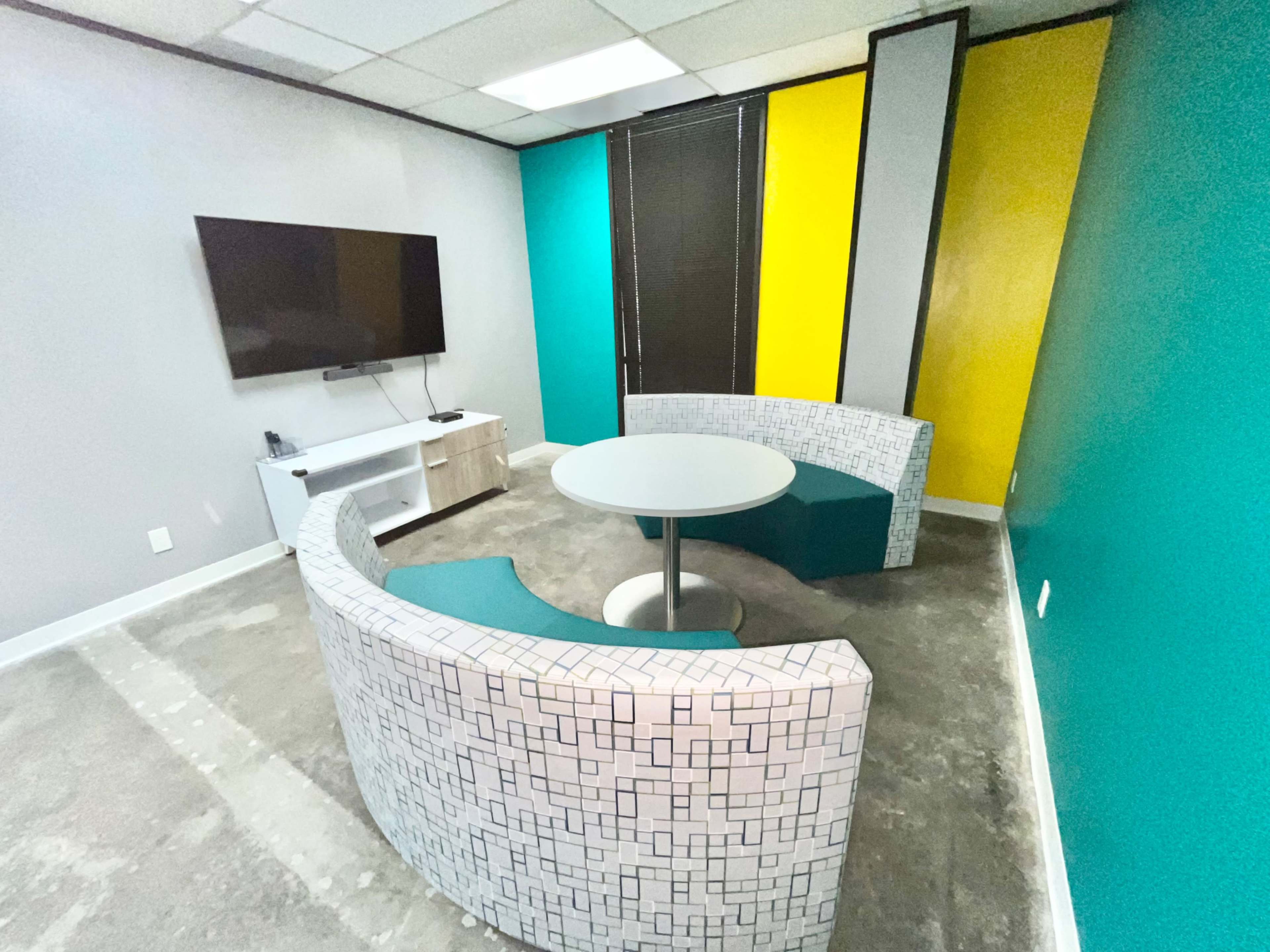 A modern meeting room features curved seating, a round table, and a television mounted on the wall, with vibrant teal and yellow accent walls.