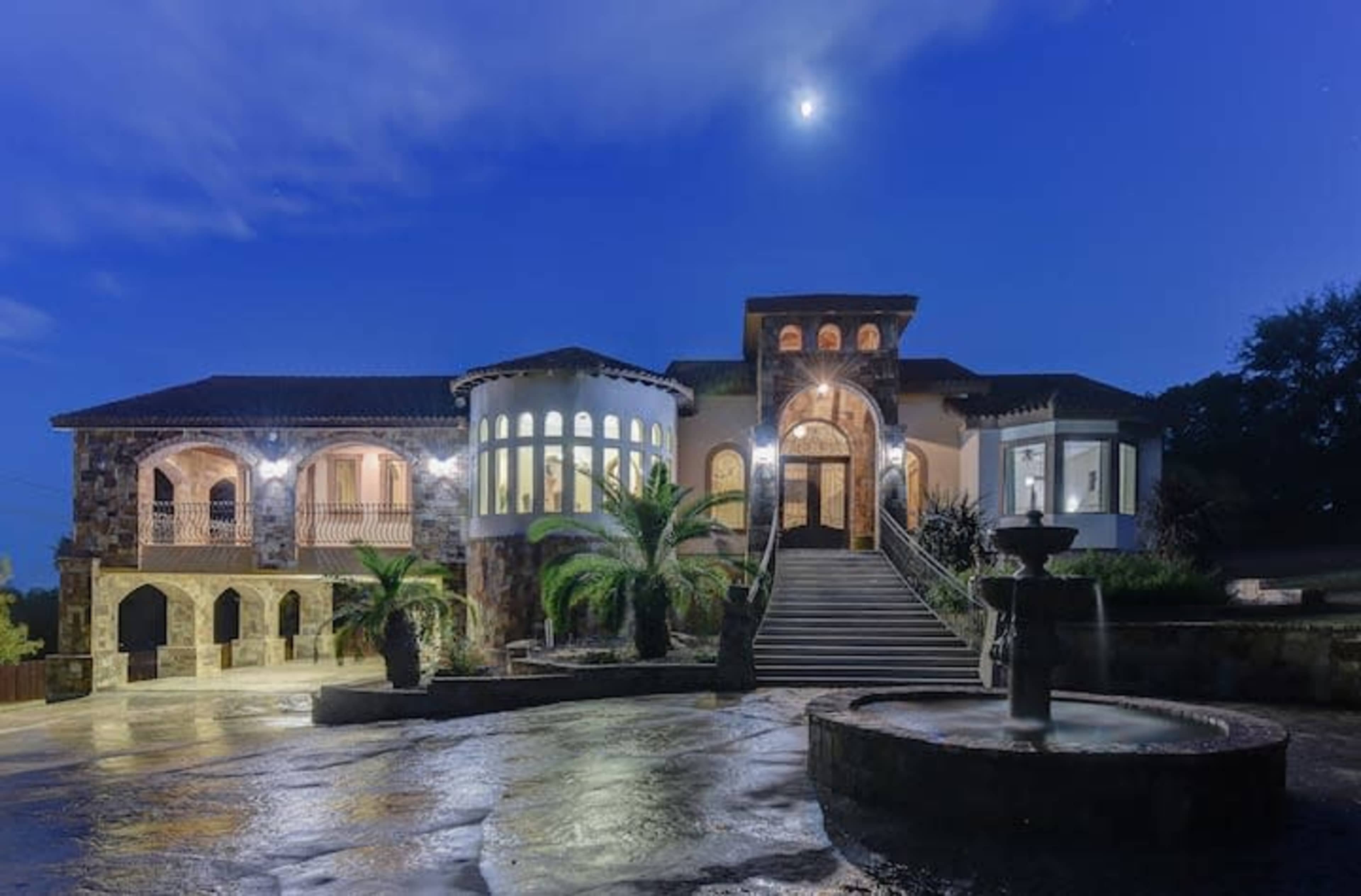 A large stone and stucco mansion with a circular turret, spacious stairway, and a fountain, illuminated under a night sky with a visible moon.