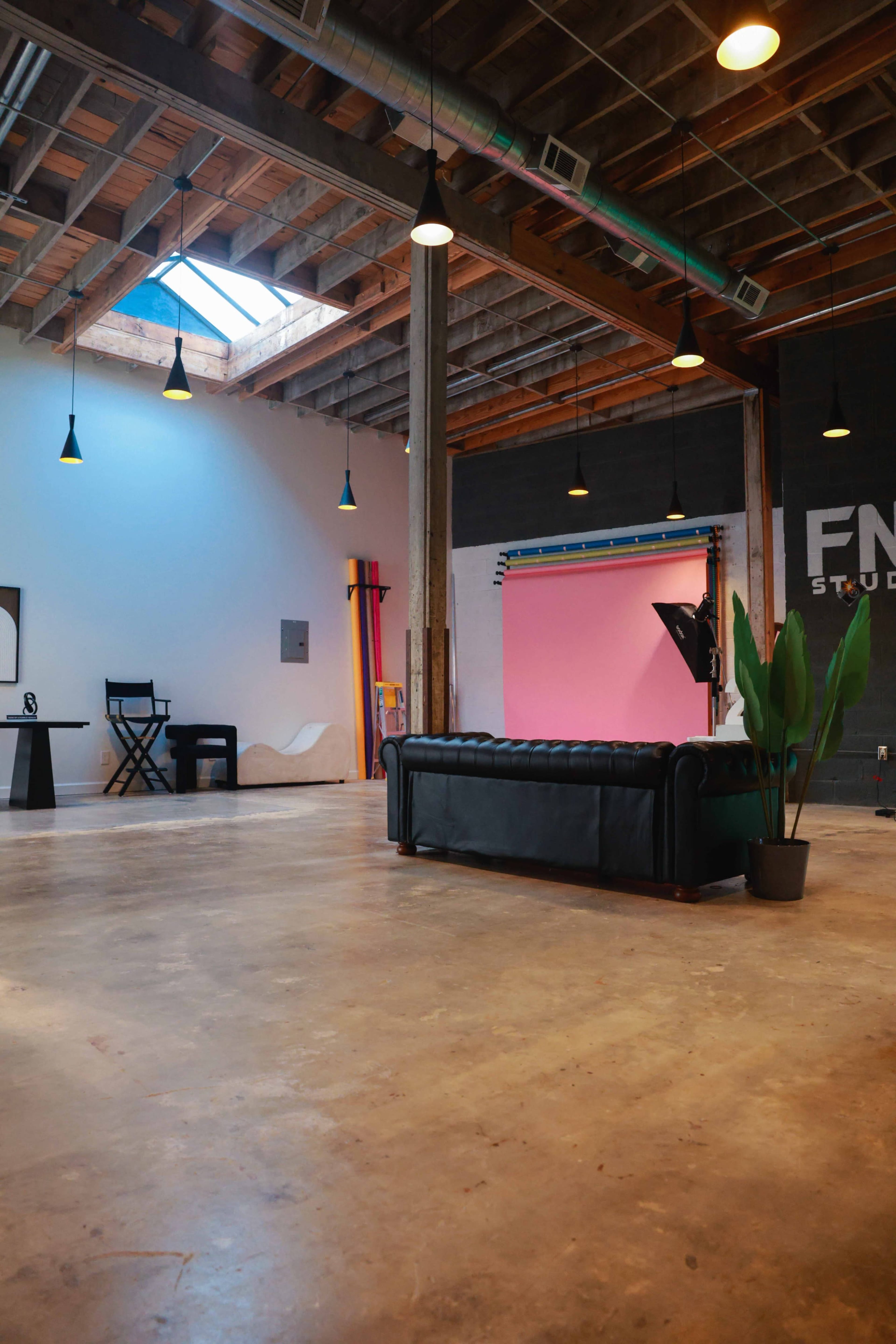 The image shows a spacious studio with a black leather couch, overhead lighting, and a colorful backdrop setup on the side.