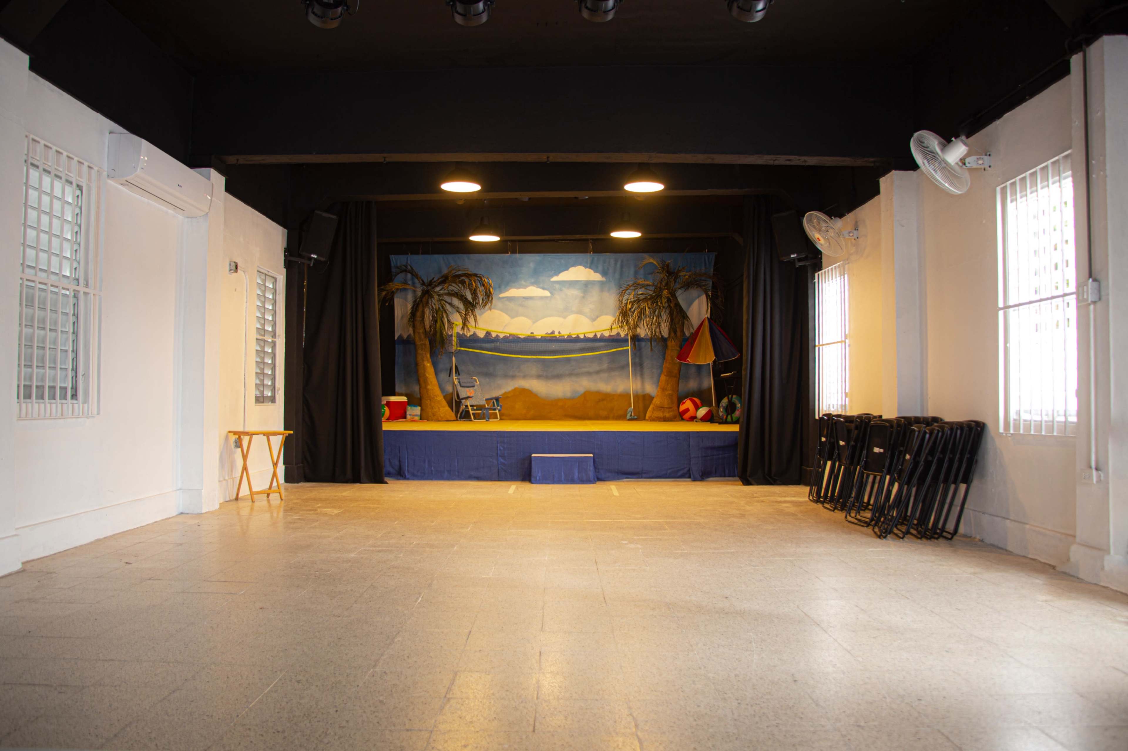 The image shows a stage set with a beach backdrop and props, including palm trees and a beach ball, in a spacious, empty room with folding chairs stacked against the wall.