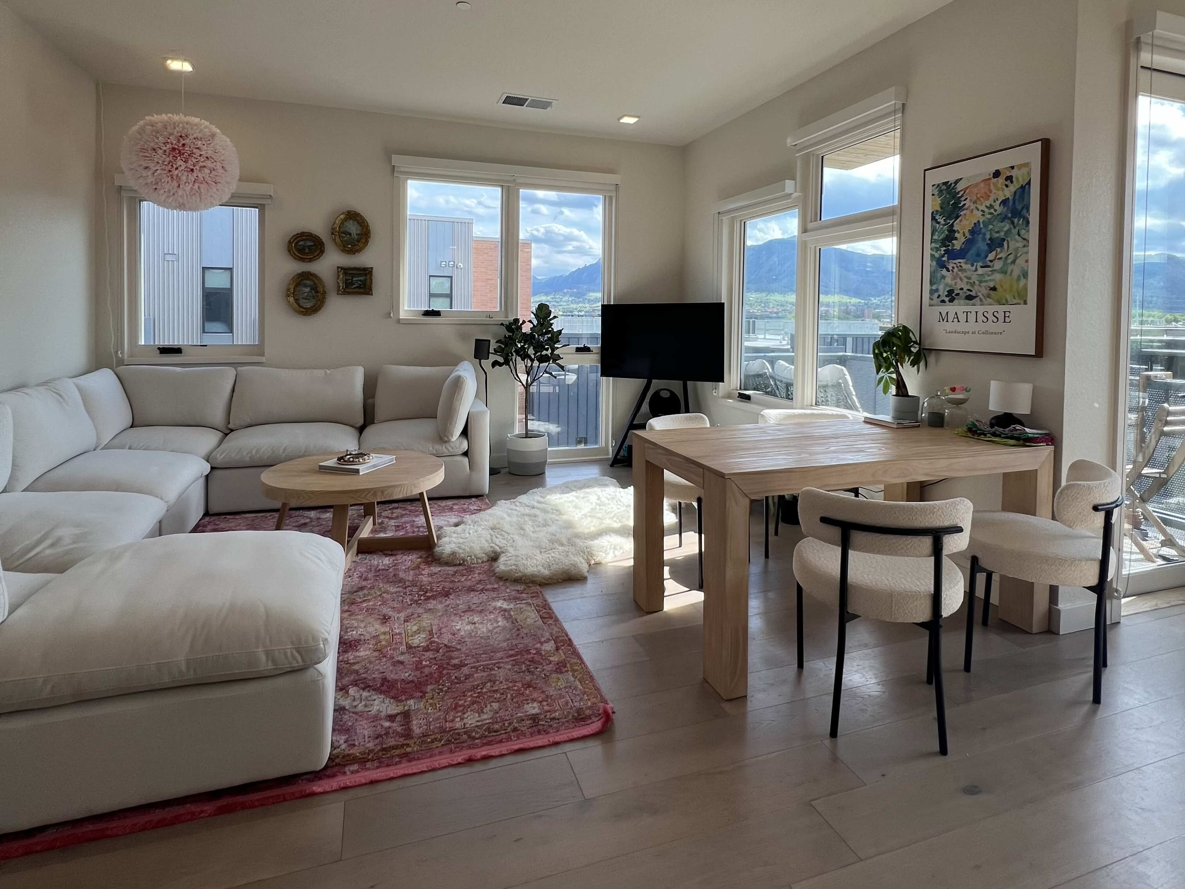 A bright living room features a white sectional couch, a wooden dining table surrounded by chairs, and large windows that offer a view of the outdoors.