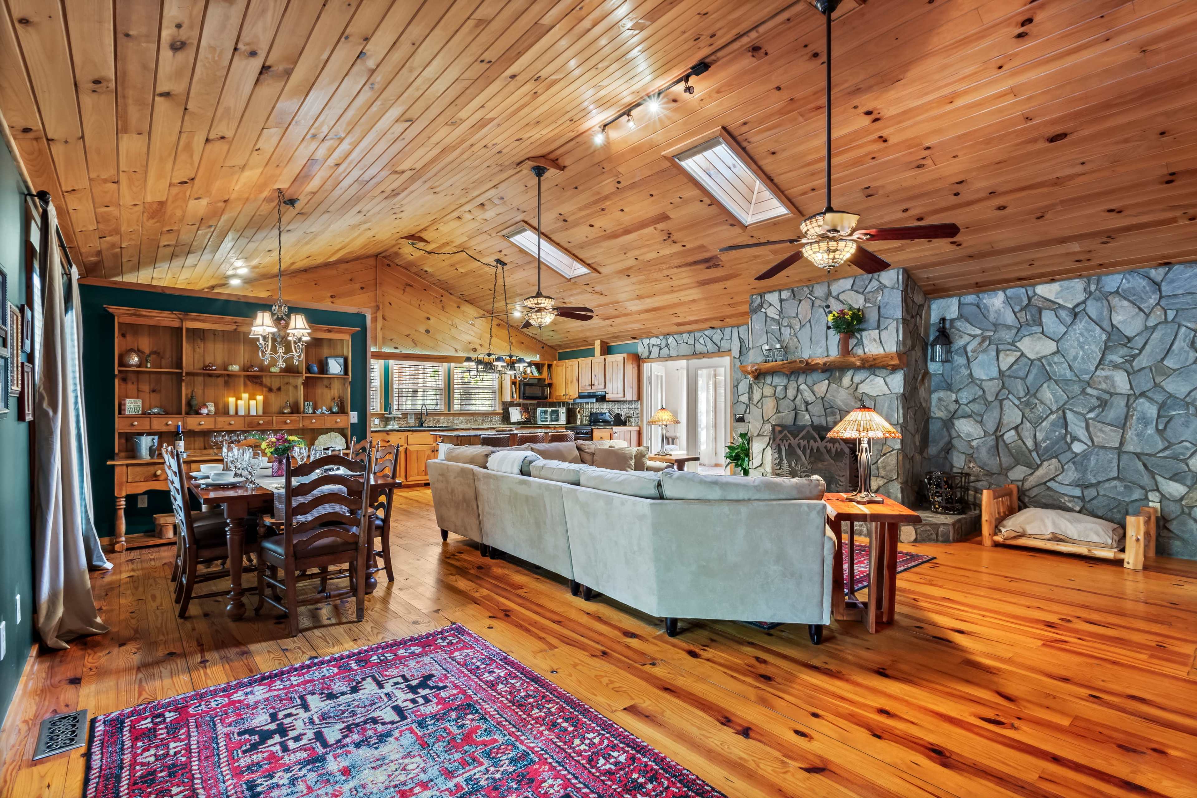 The image displays a spacious living area with a wooden ceiling, stone fireplace, and a blend of dining and seating arrangements.