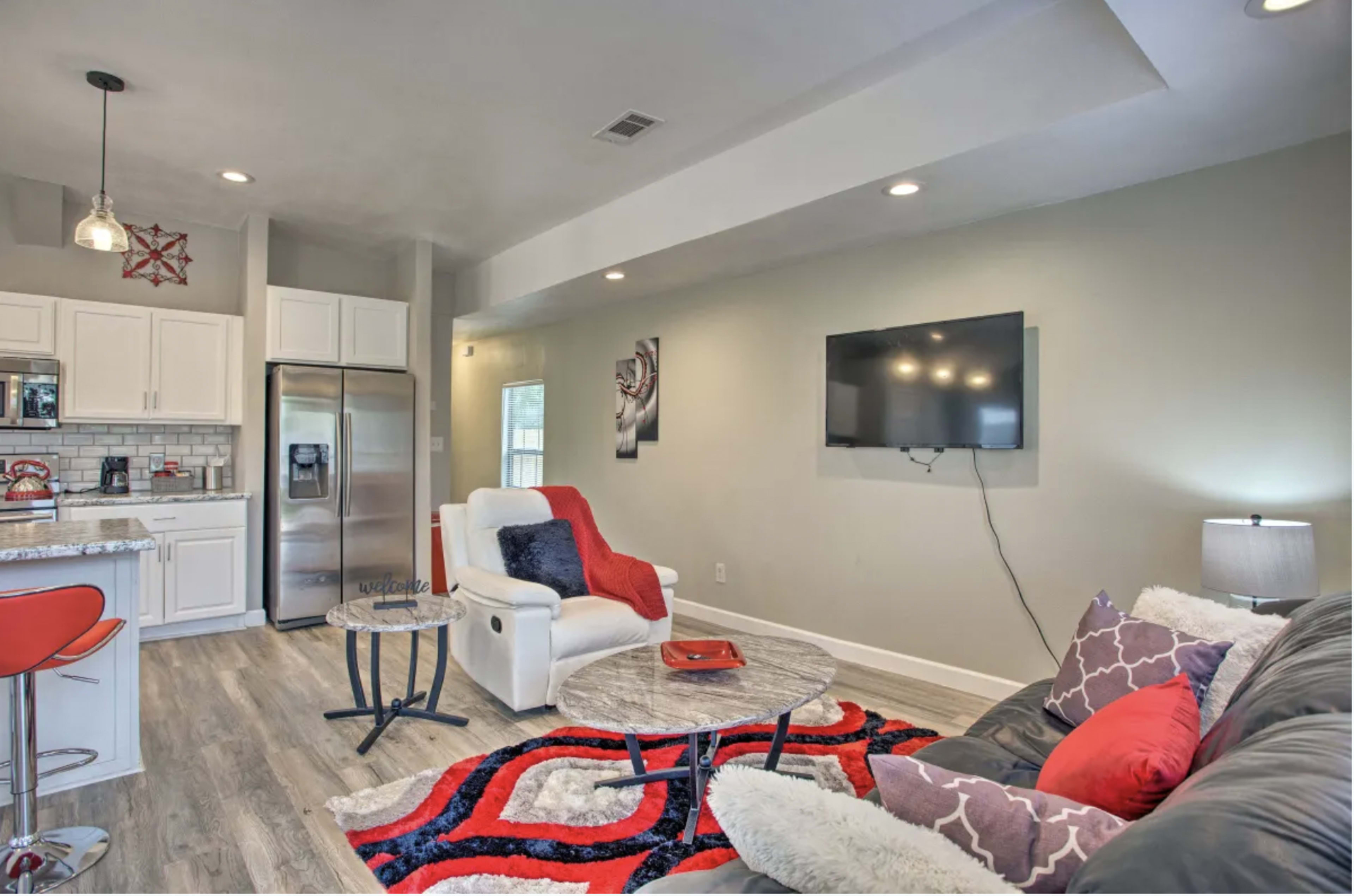 A modern living area featuring a gray sofa with colorful cushions, a coffee table, a wall-mounted television, and a kitchenette with stainless steel appliances.
