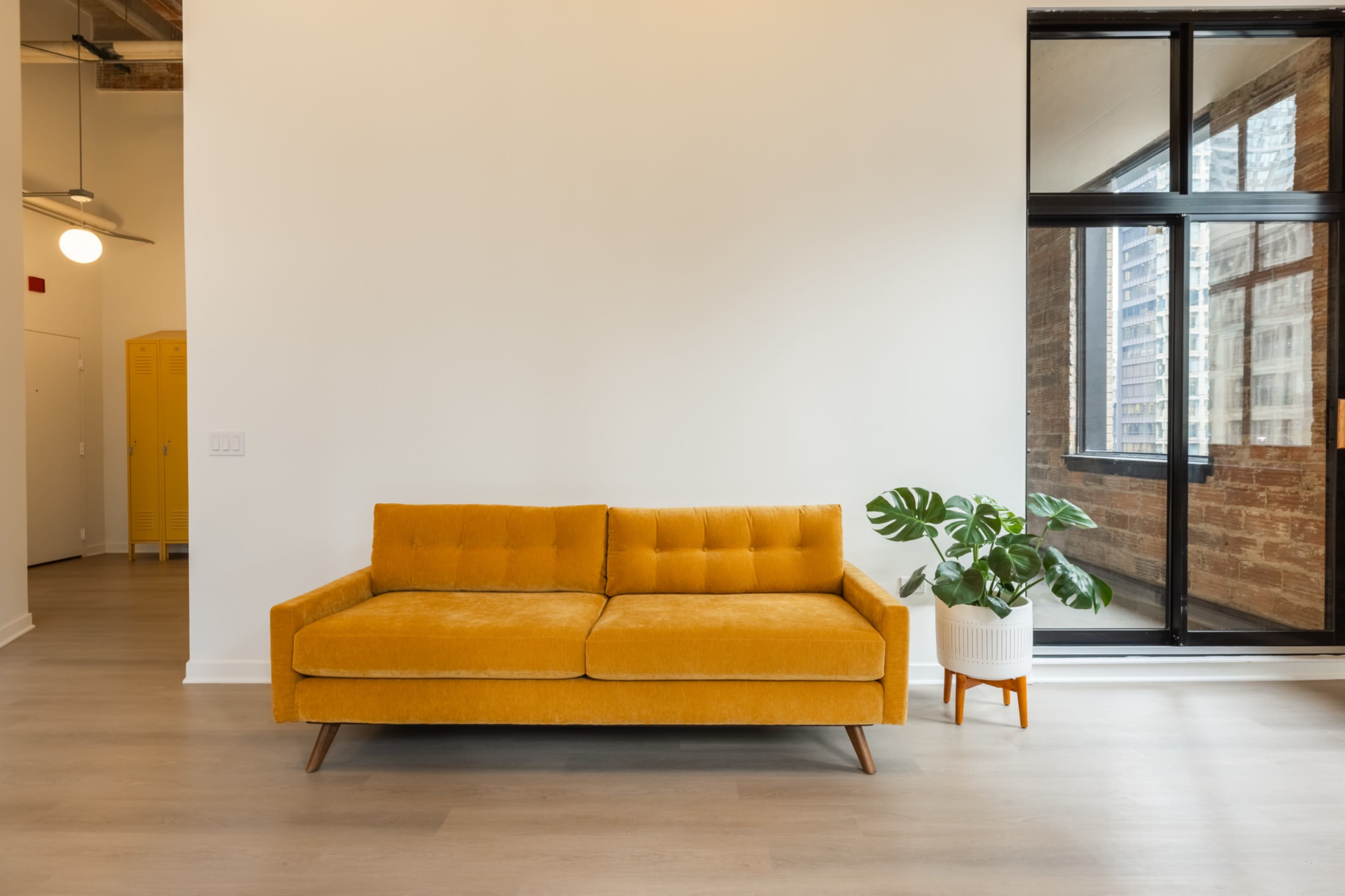 The image shows a yellow couch in a minimalist living space with a large window and a potted plant nearby.