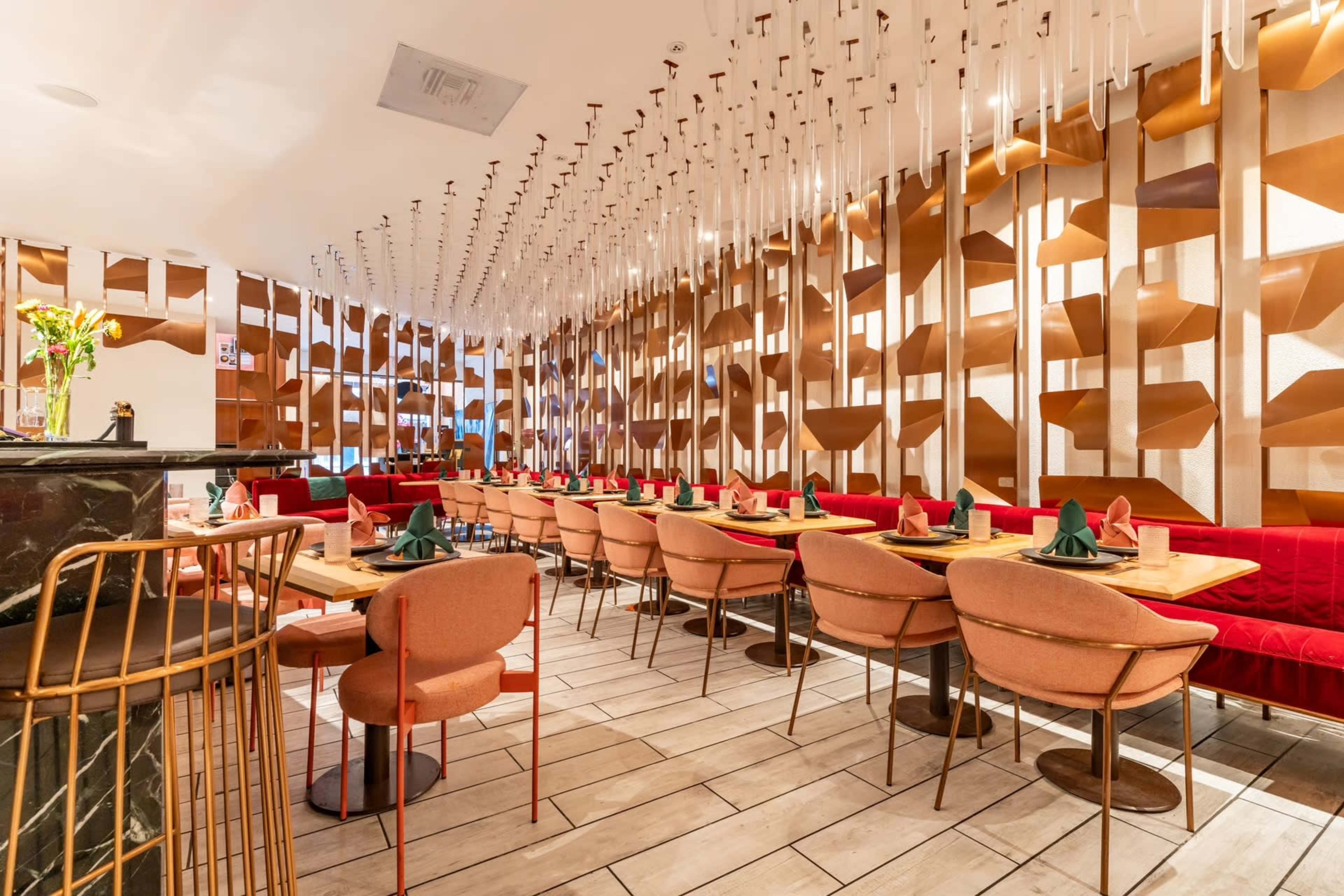 The image shows a modern restaurant interior with rows of red upholstered chairs and wooden tables, featuring decorative lighting and textured wall panels.