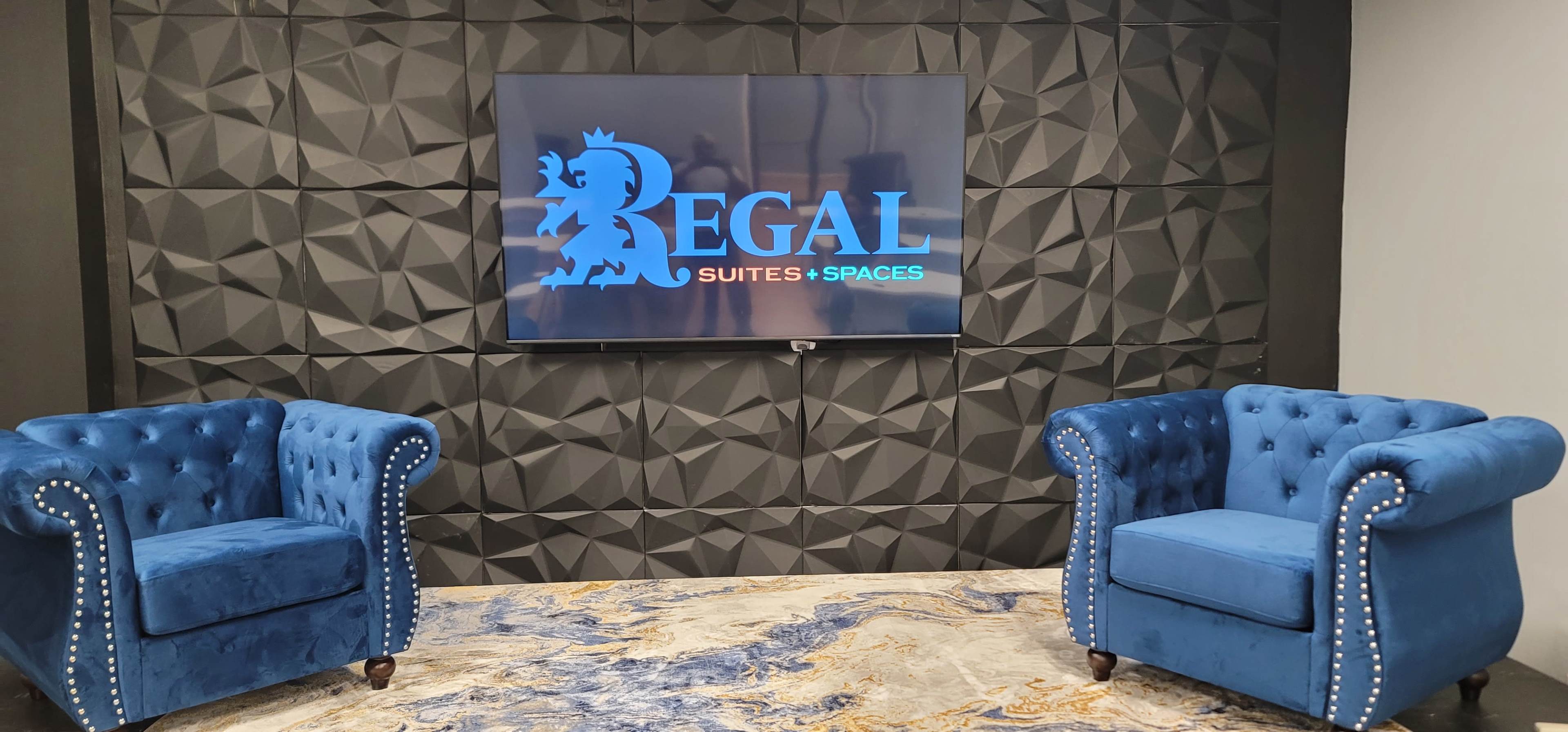 Two blue tufted armchairs facing a wall-mounted television displaying the logo "REGAL Suites & Spaces" against a textured black background.