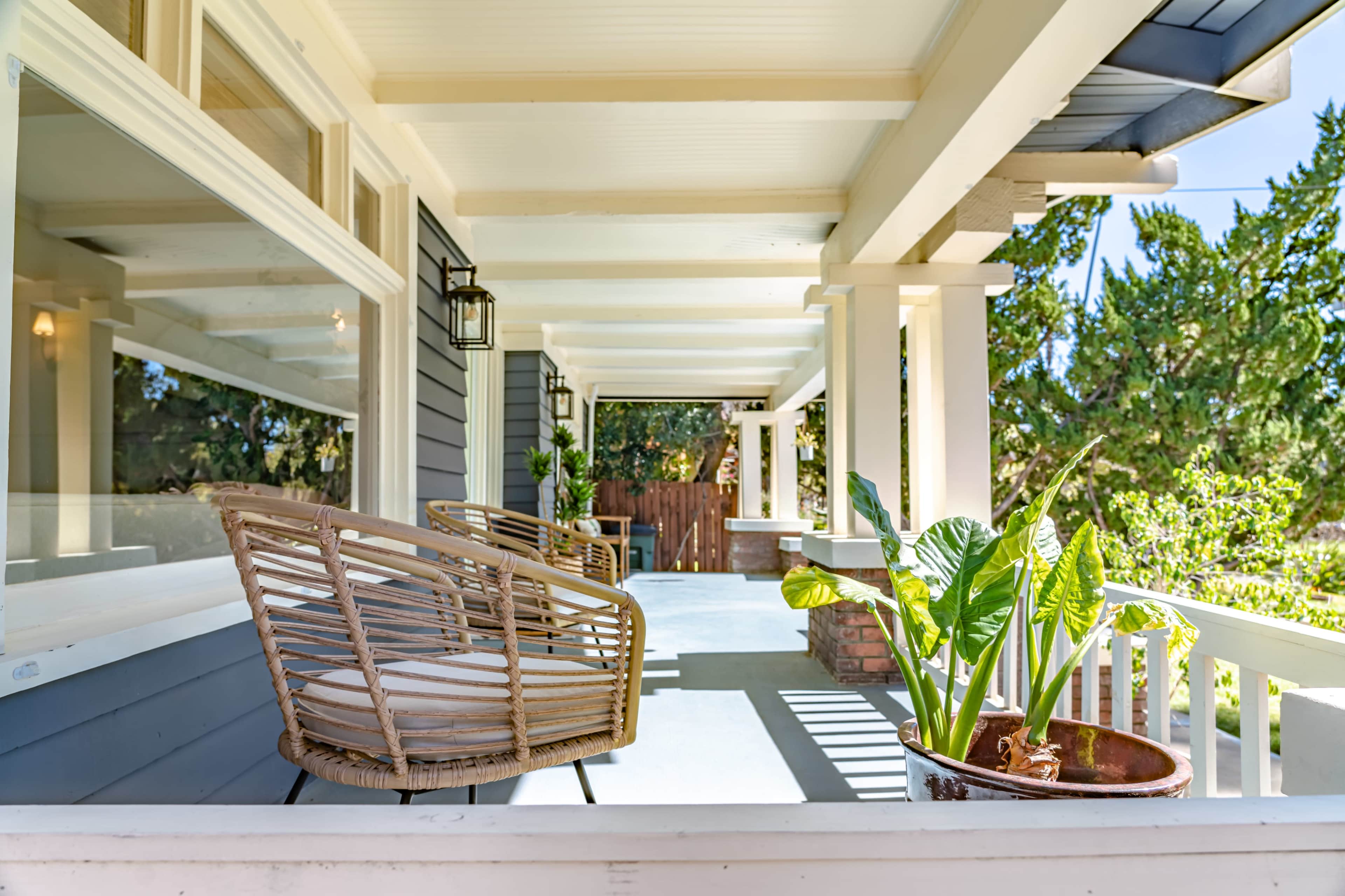 A porch featuring two wicker chairs and a potted plant under the shade of wooden columns.