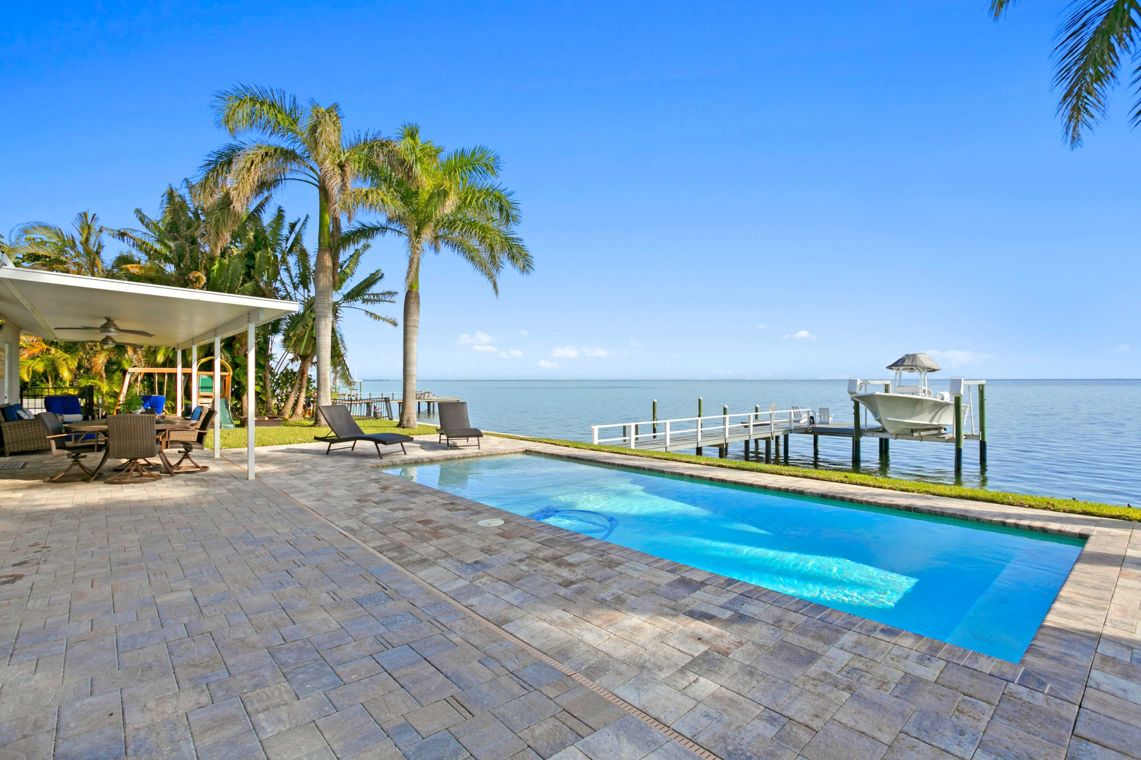 The image shows a patio with a swimming pool and a view of the waterfront, featuring palm trees and a dock extending into the water.