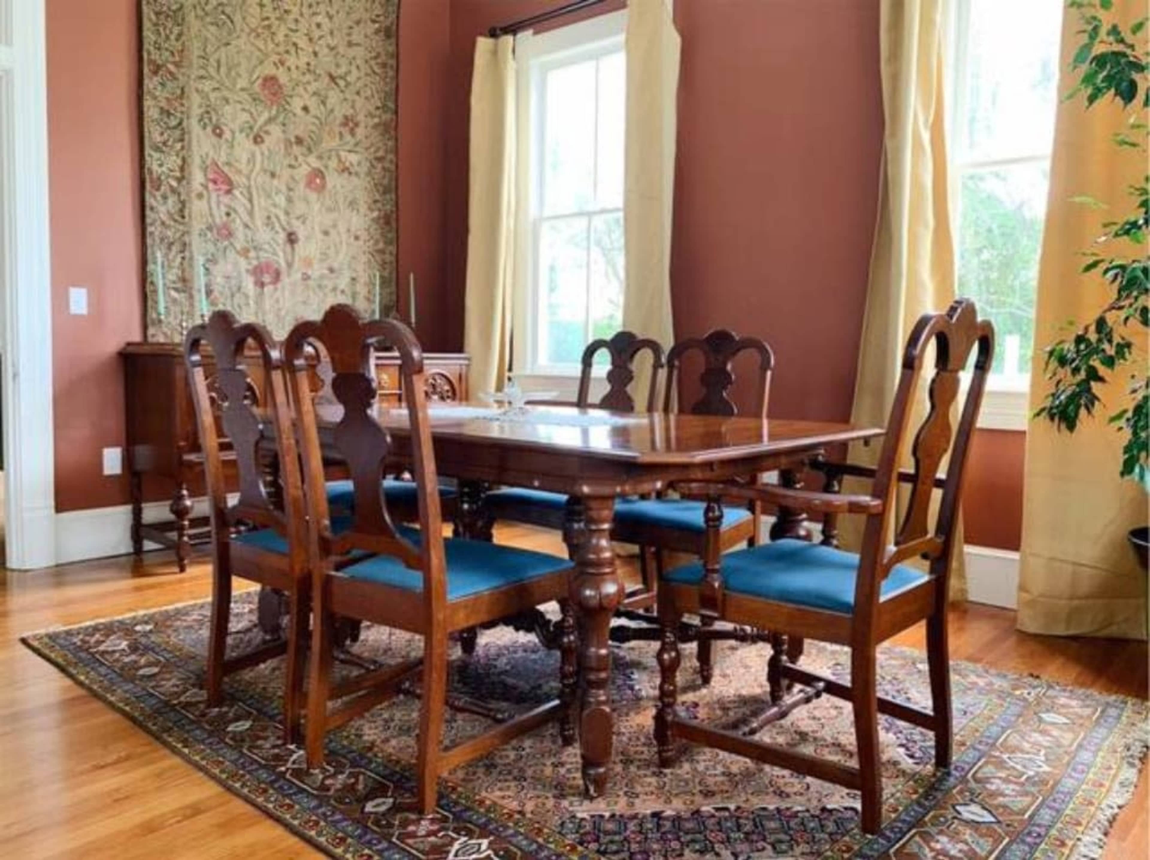 A wooden dining table surrounded by six chairs is set in a room with large windows and a decorative rug.