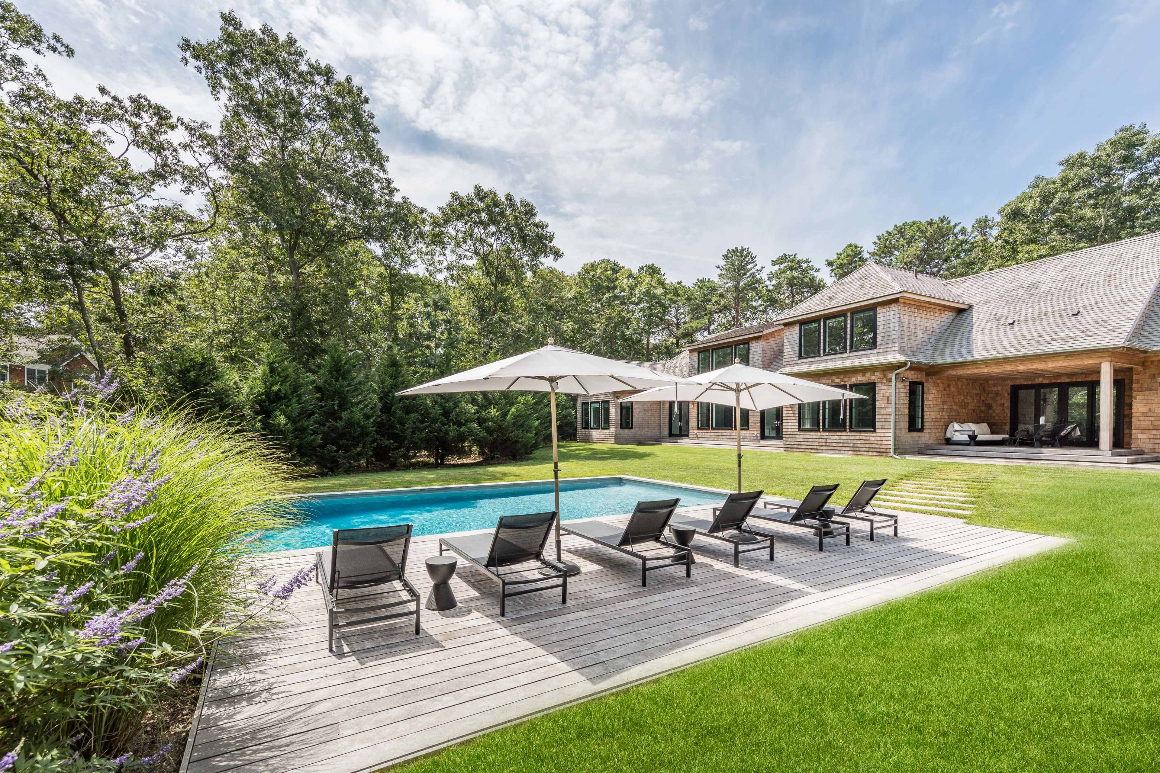 A landscaped yard features a swimming pool with lounge chairs and umbrellas beside a residential home surrounded by trees.