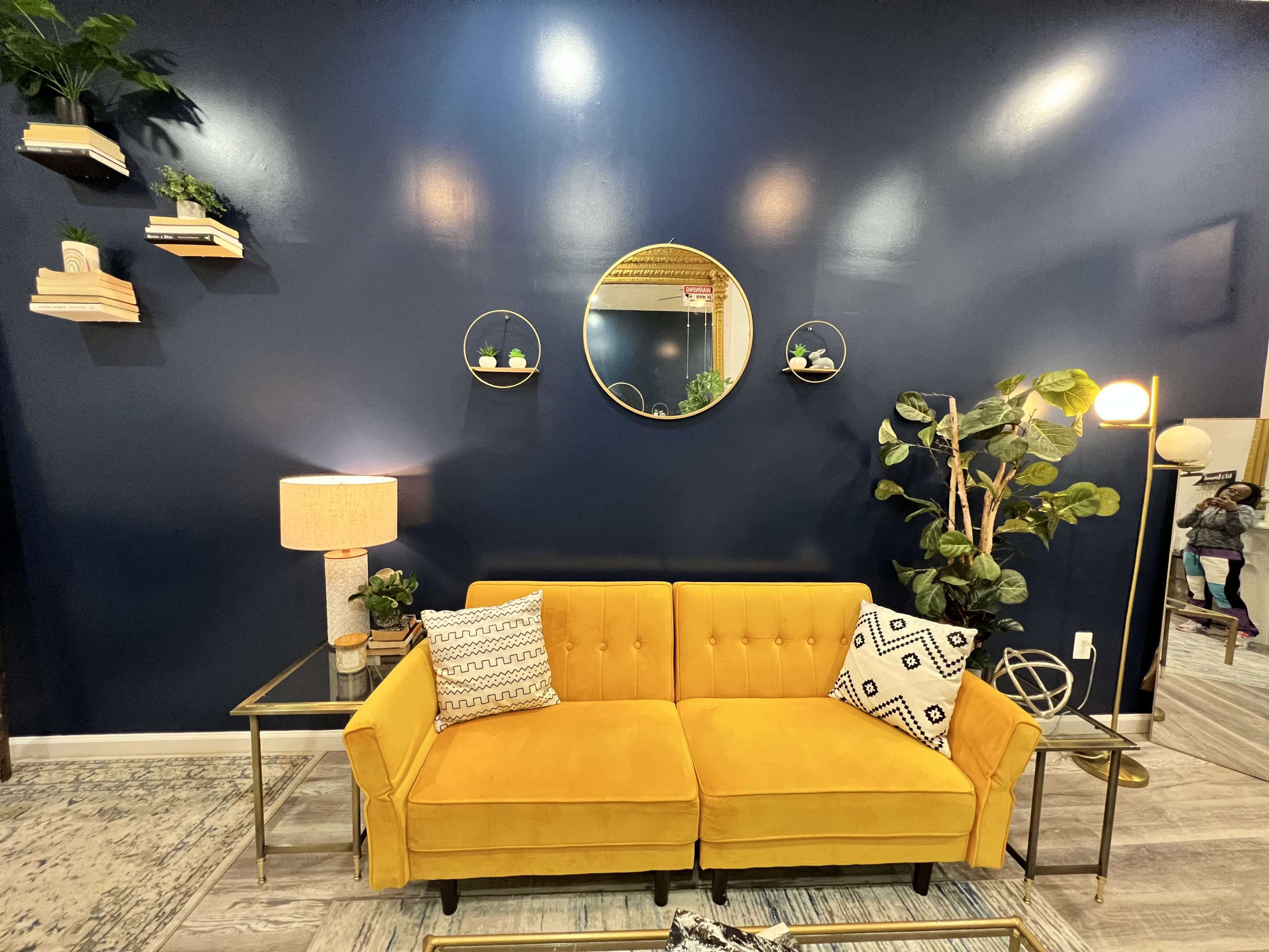 The image shows a cozy living room with a yellow couch, a round mirror on a dark blue wall, and various plants displayed on shelves.