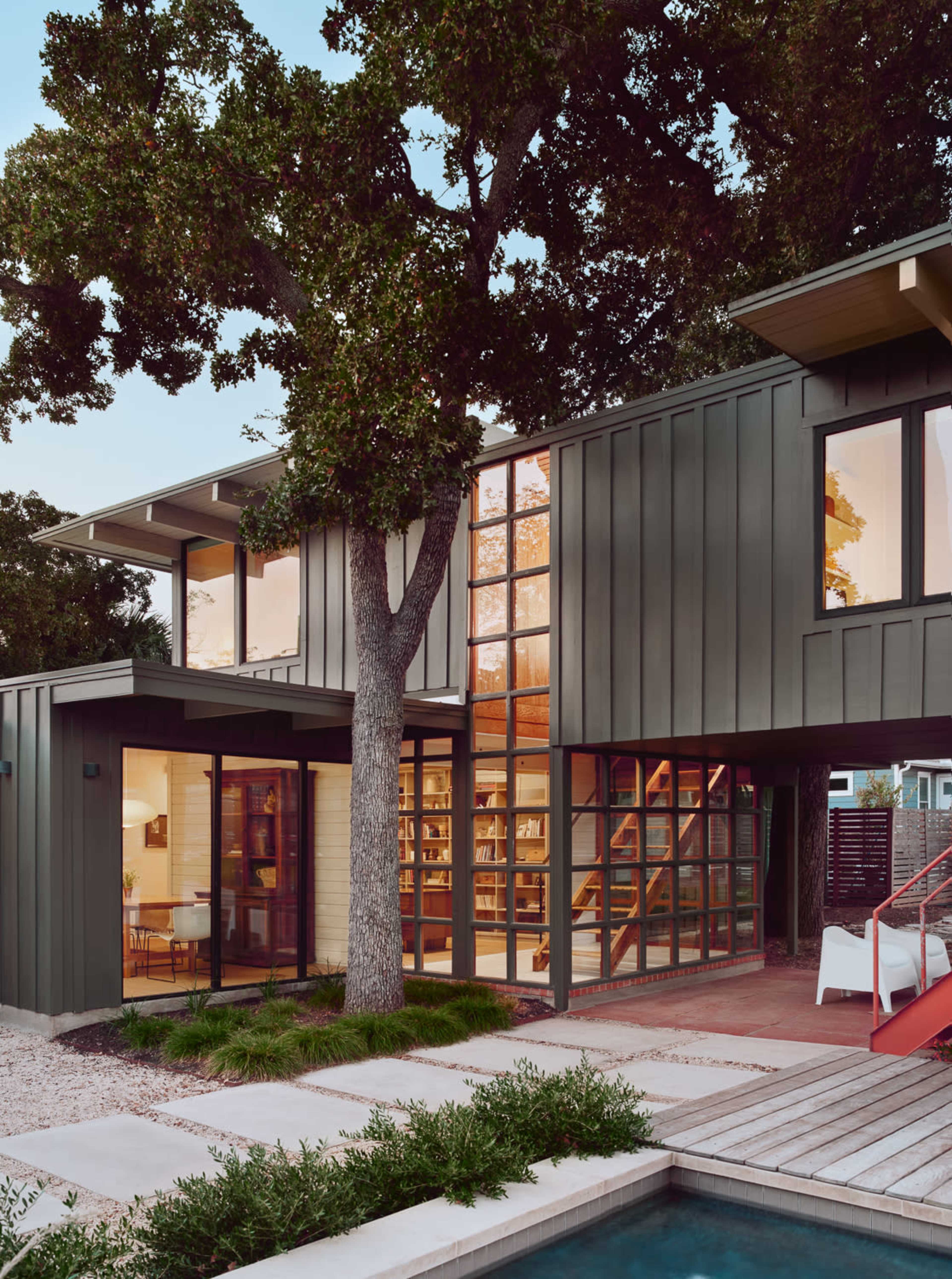 A modern two-story house features large windows, a tree beside it, and a wooden deck near a swimming pool.