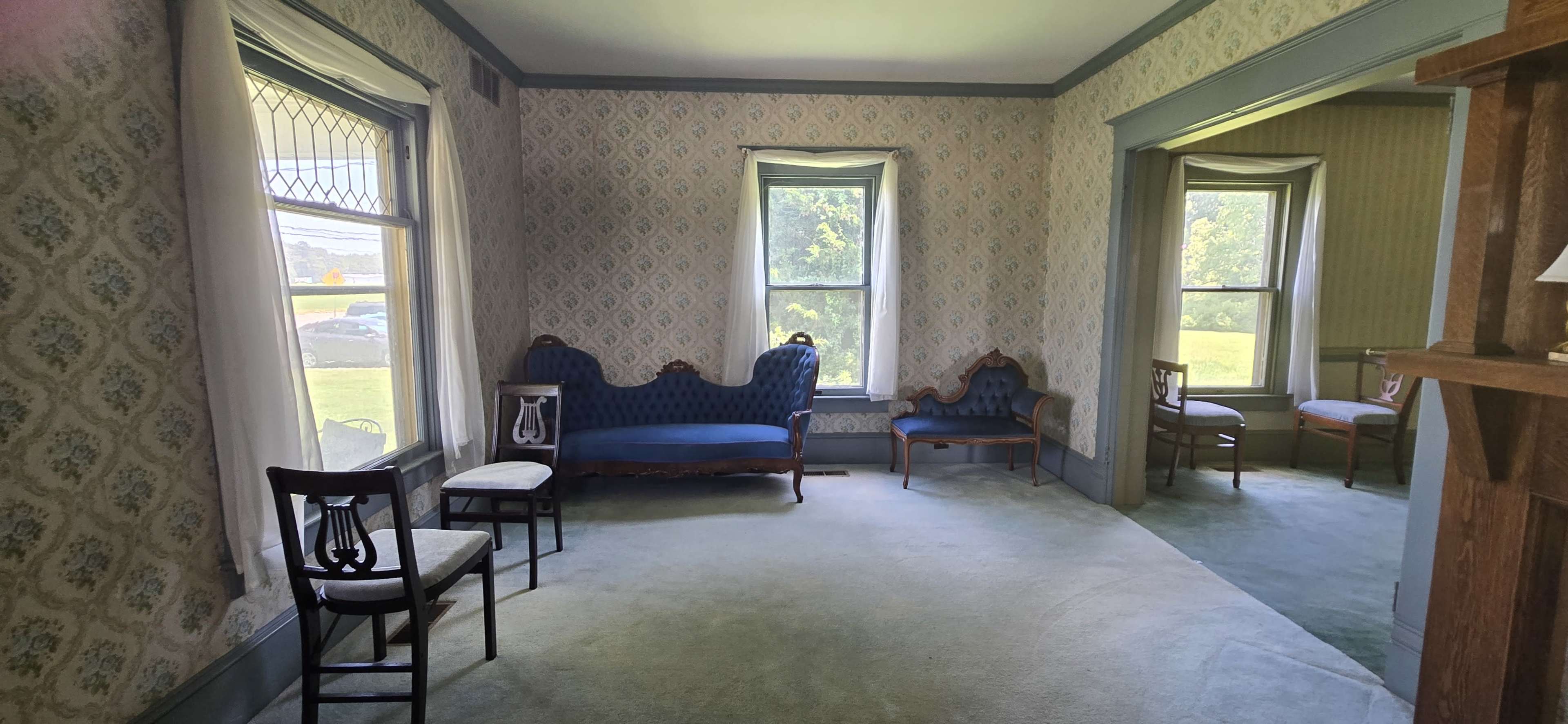 The image depicts a room furnished with blue upholstered seating, several chairs, and windows that allow light to illuminate the space.