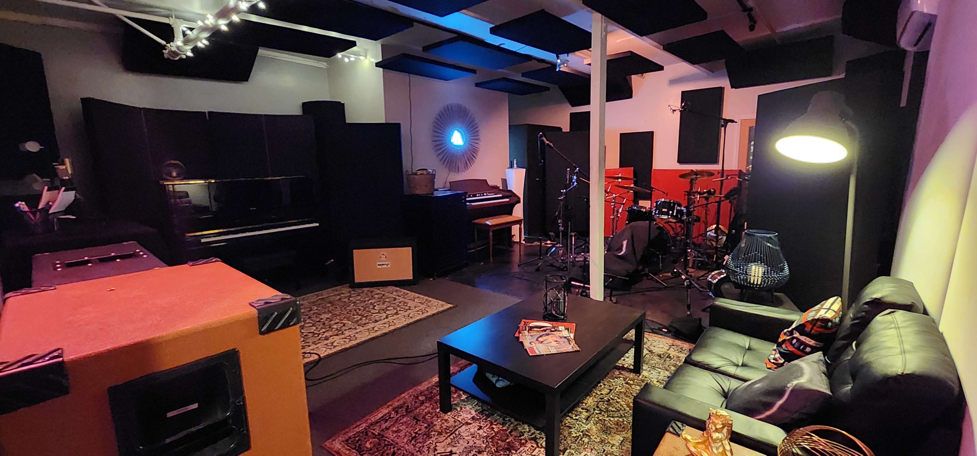 The image shows a soundproof music studio featuring a piano, drum set, and a cozy seating area with a coffee table.