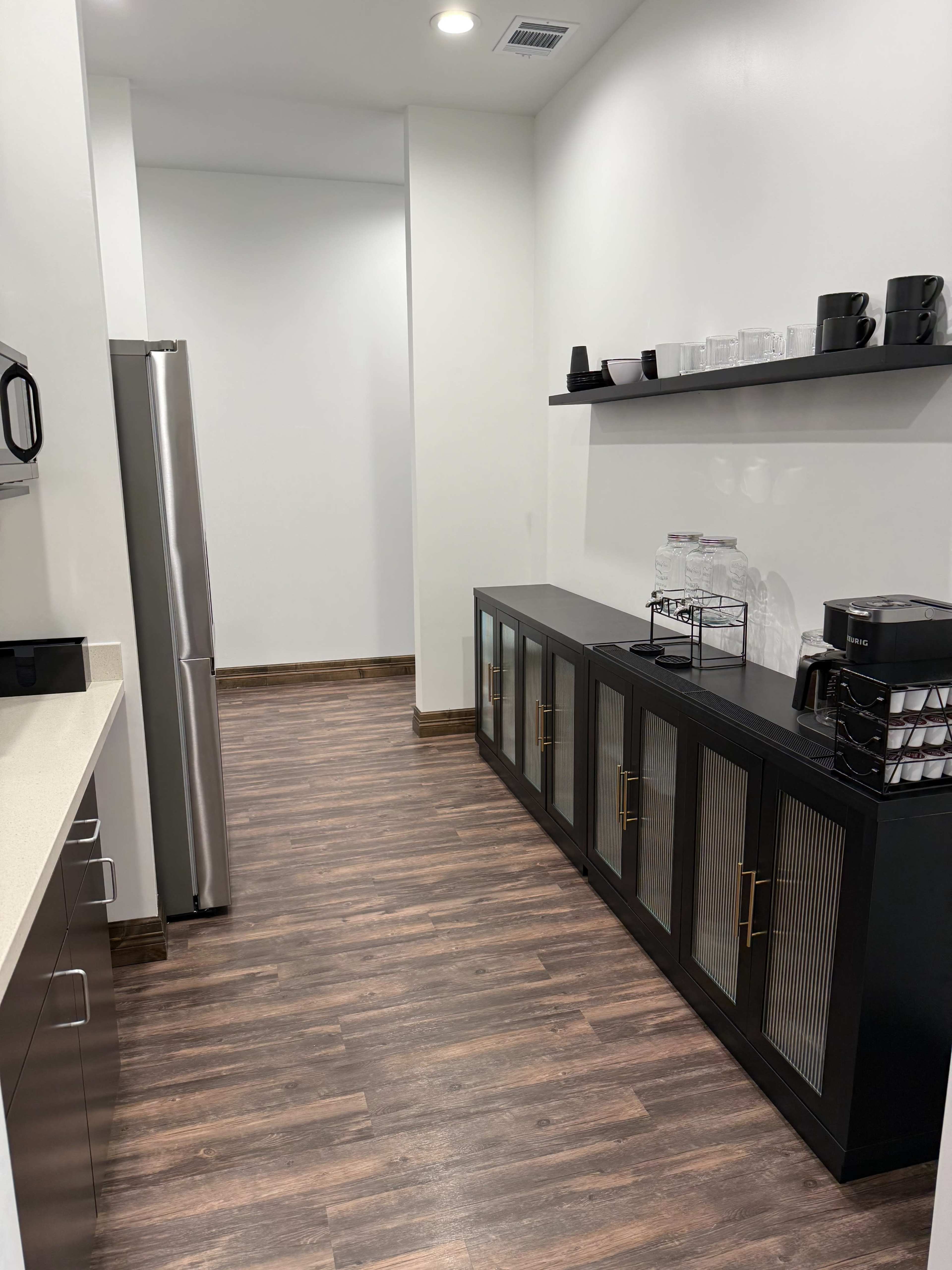 The image shows a modern kitchen area with a stainless steel refrigerator, a quartz countertop, and a dark cabinet containing glassware and utensils along one wall.