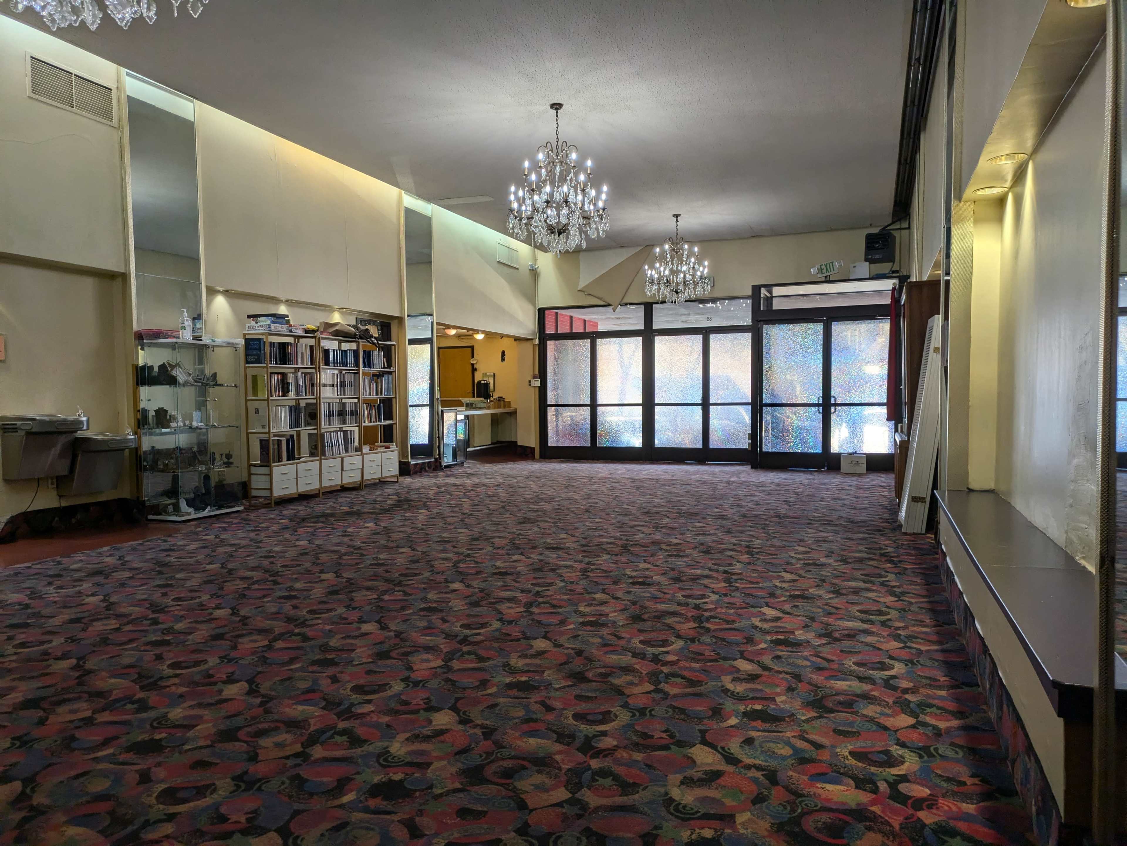 The image shows an empty indoor space with a patterned carpet, large glass doors at the back, and various light fixtures hanging from the ceiling.
