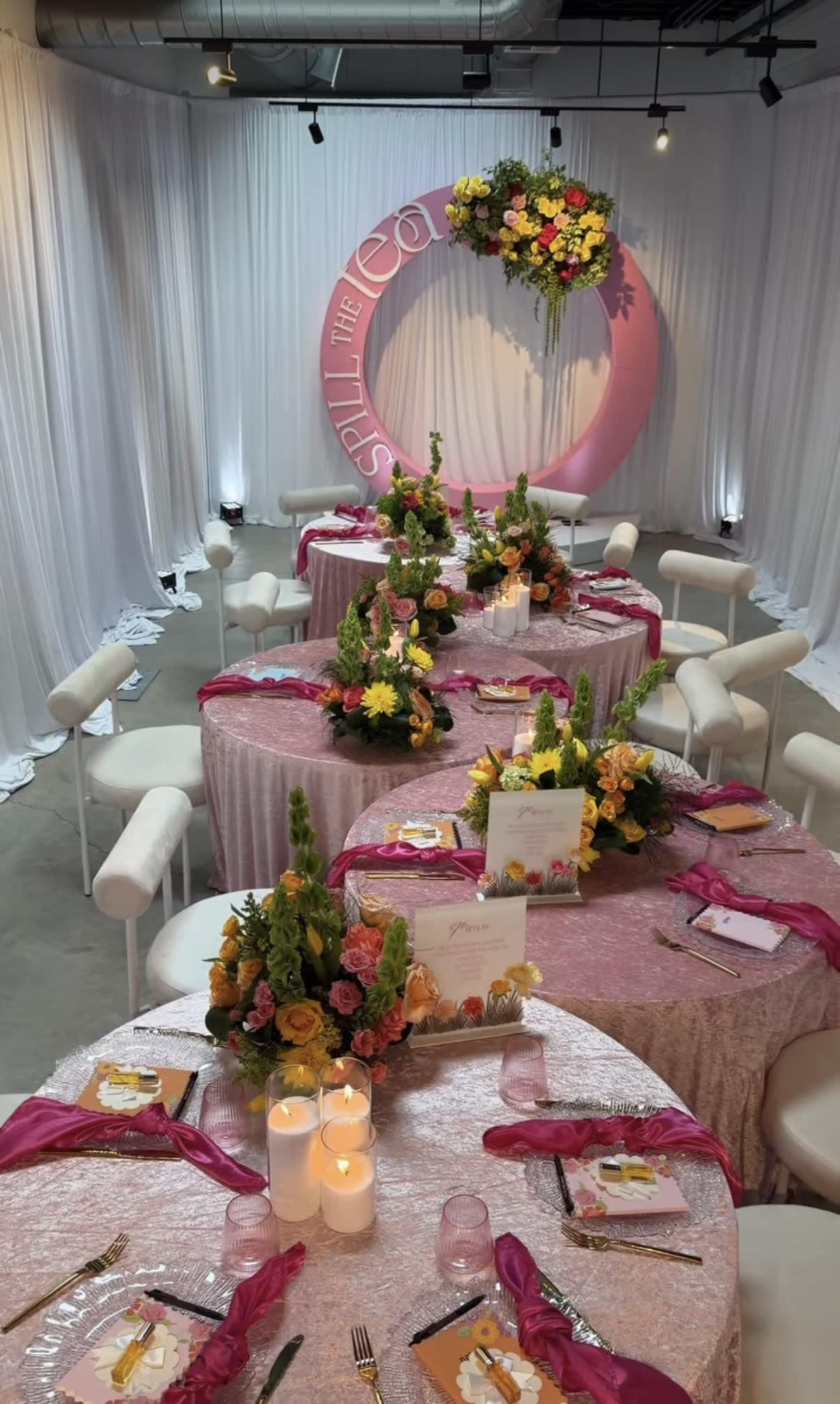 The image shows a beautifully arranged event space with circular tables dressed in light pink tablecloths, adorned with floral centerpieces and candles, and a decorative backdrop featuring a large circular sign.