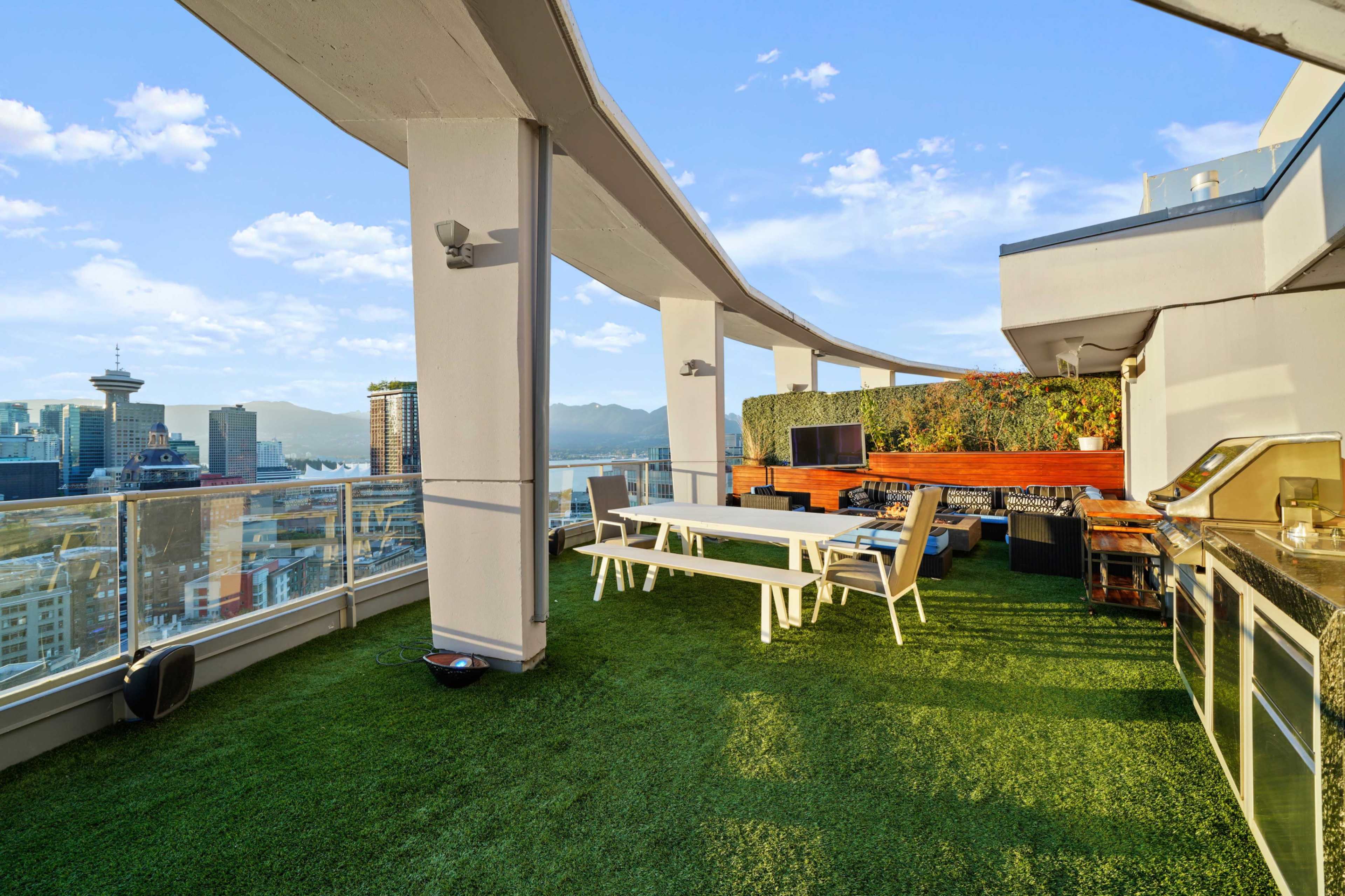 The image shows a rooftop terrace featuring a grassy area, a dining table, an outdoor kitchen, and a panoramic view of a city skyline with mountains in the background.