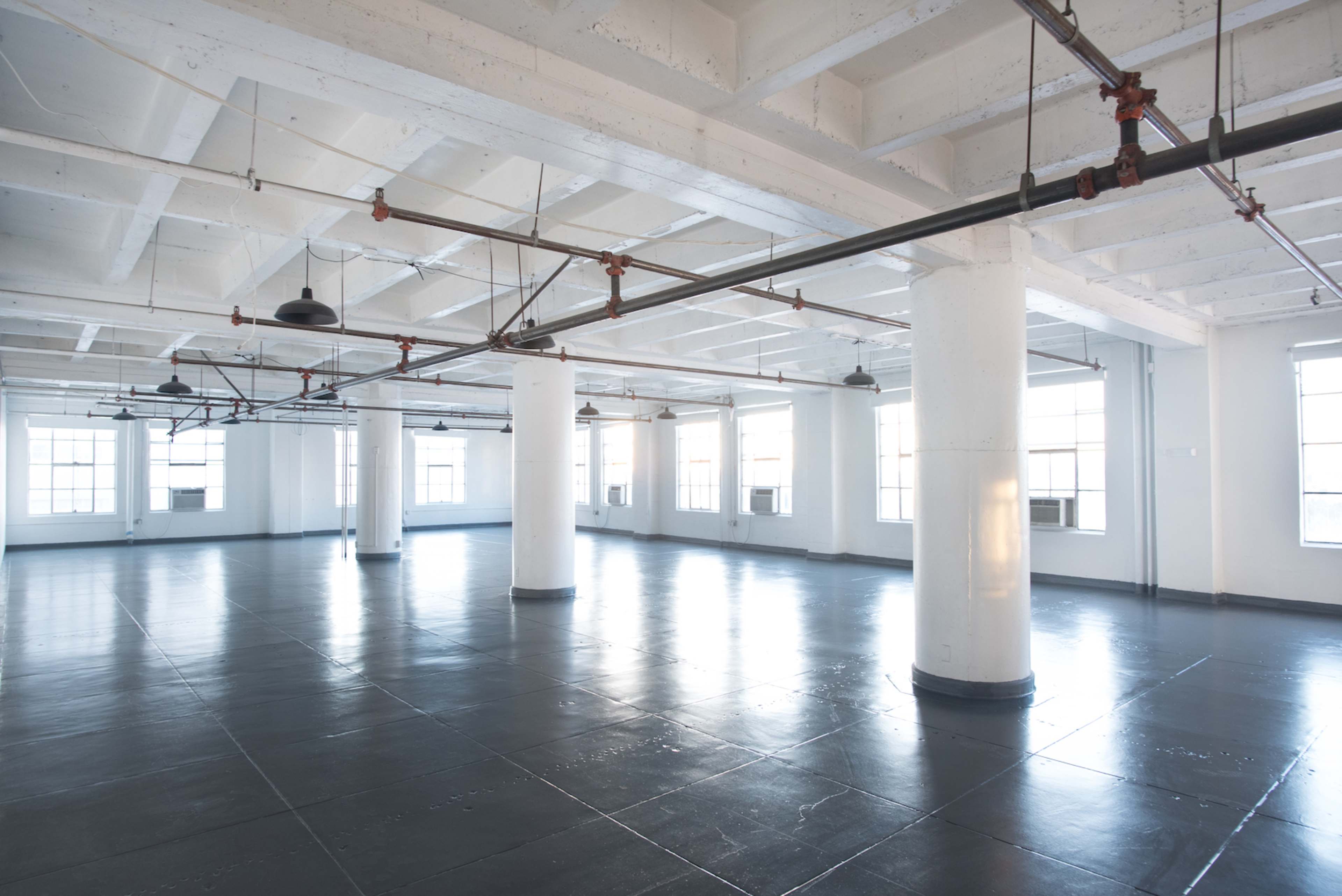 A spacious, empty industrial room with polished black floors and beams, featuring white pillars and large windows.