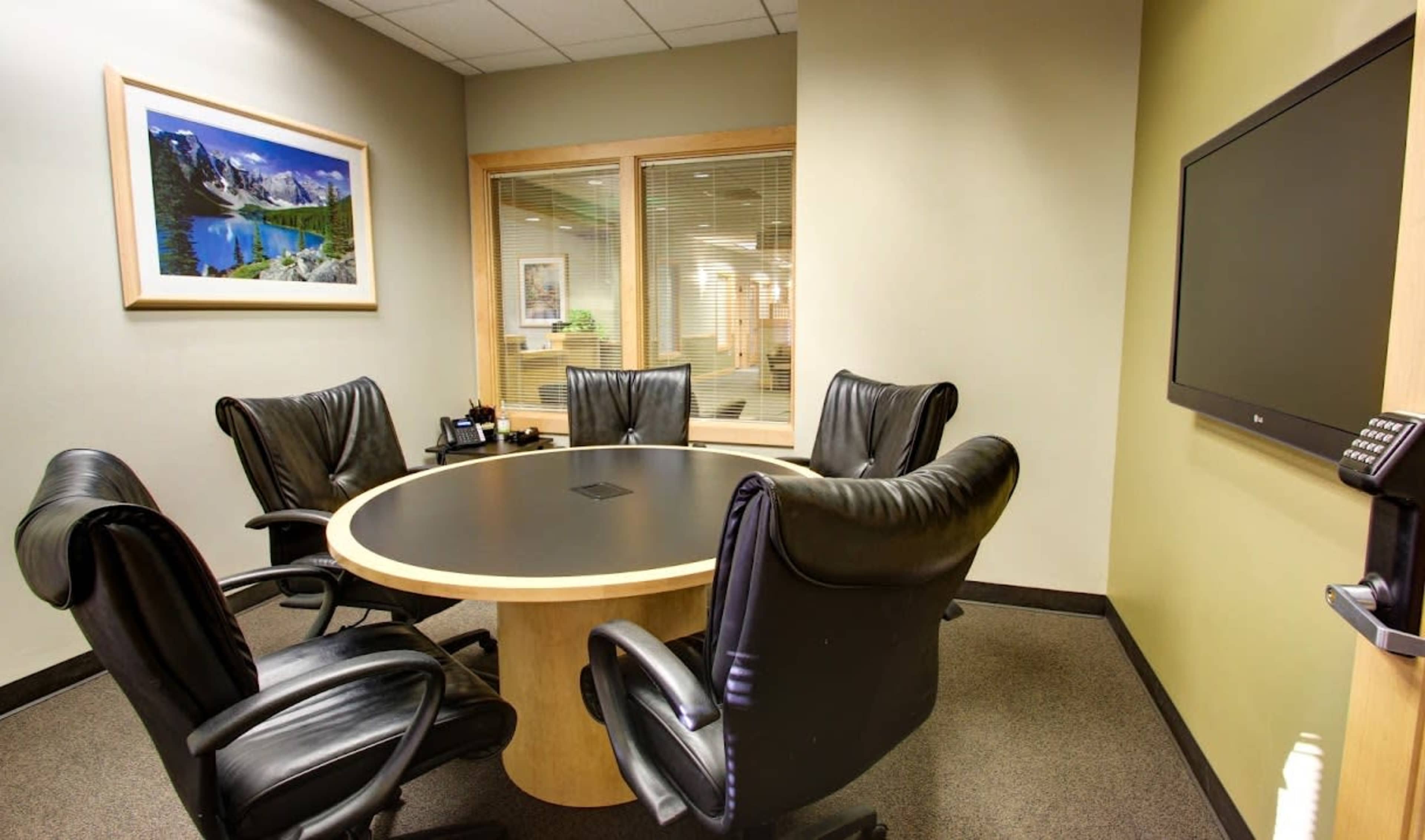 A conference room is furnished with a round table and four black leather chairs, with a television and a landscape photograph on the wall.