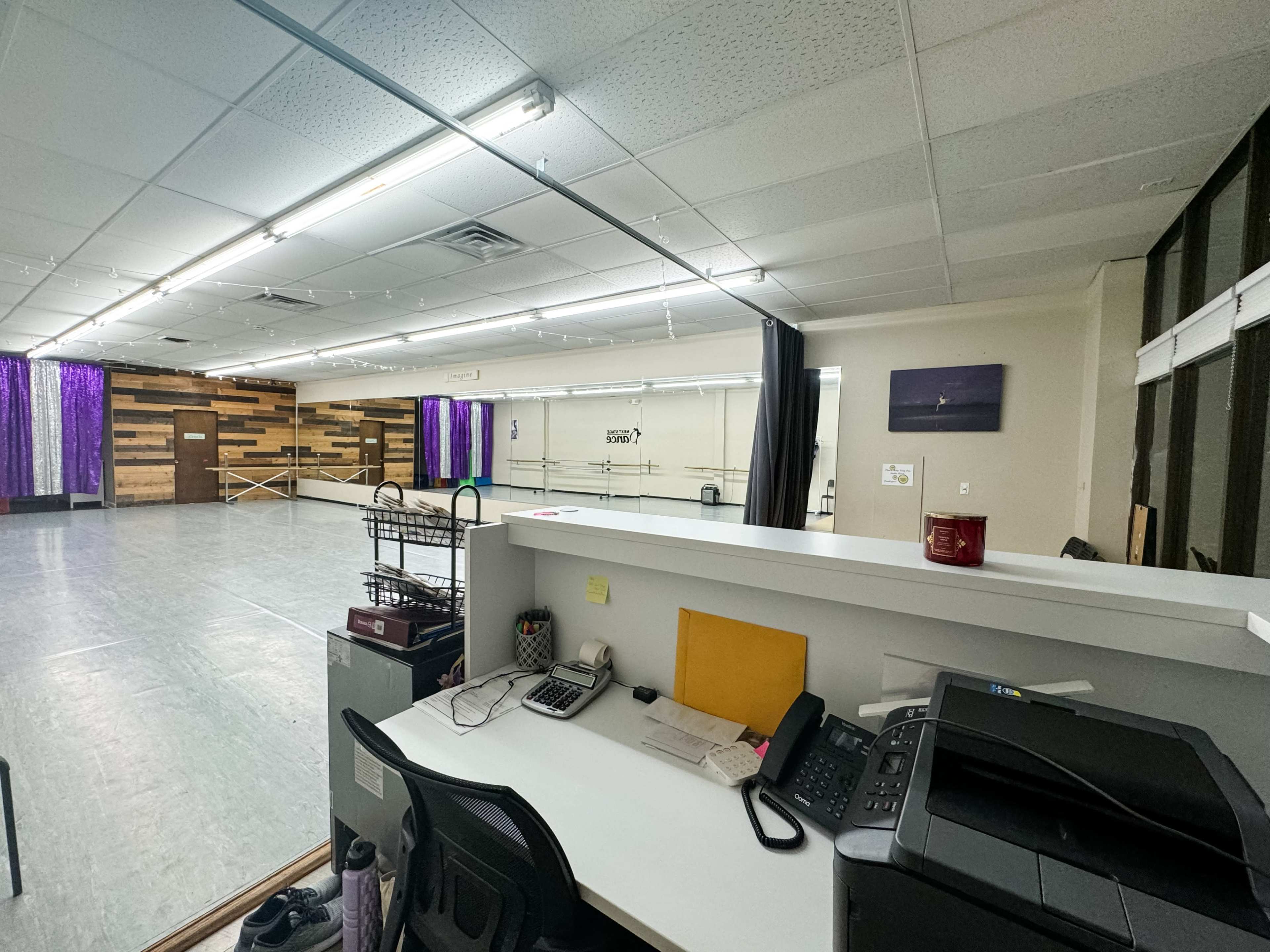 The image shows a spacious dance studio with a reception area featuring a desk, printer, and chair, and mirrors along the wall.
