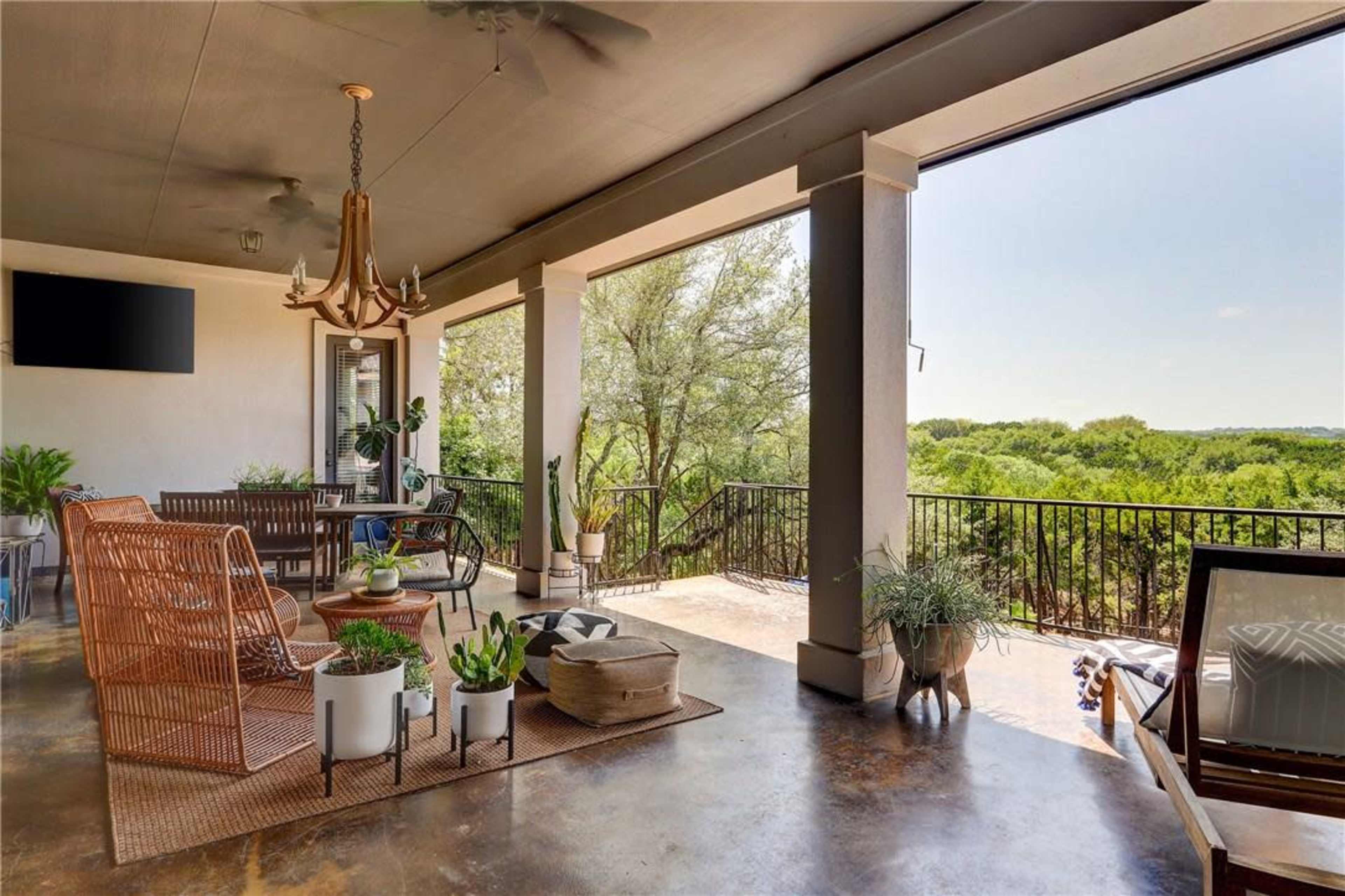 A covered patio features a seating area with wicker furniture, a chandelier, and potted plants, overlooking a green landscape.