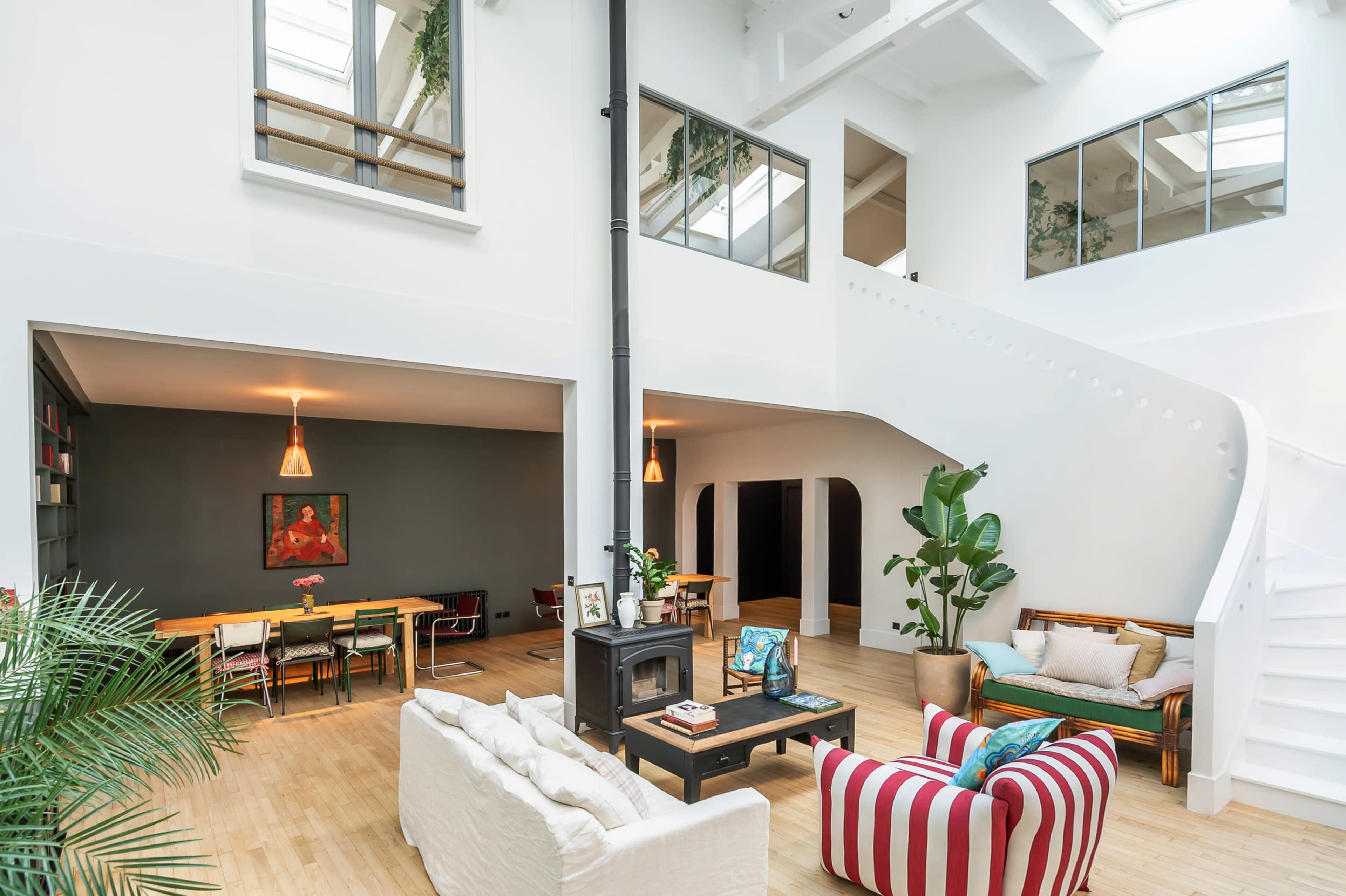 The image shows a spacious, modern interior featuring a living area with a white sofa and a colorful striped armchair, a dining area with a table, and a staircase leading to an upper level.