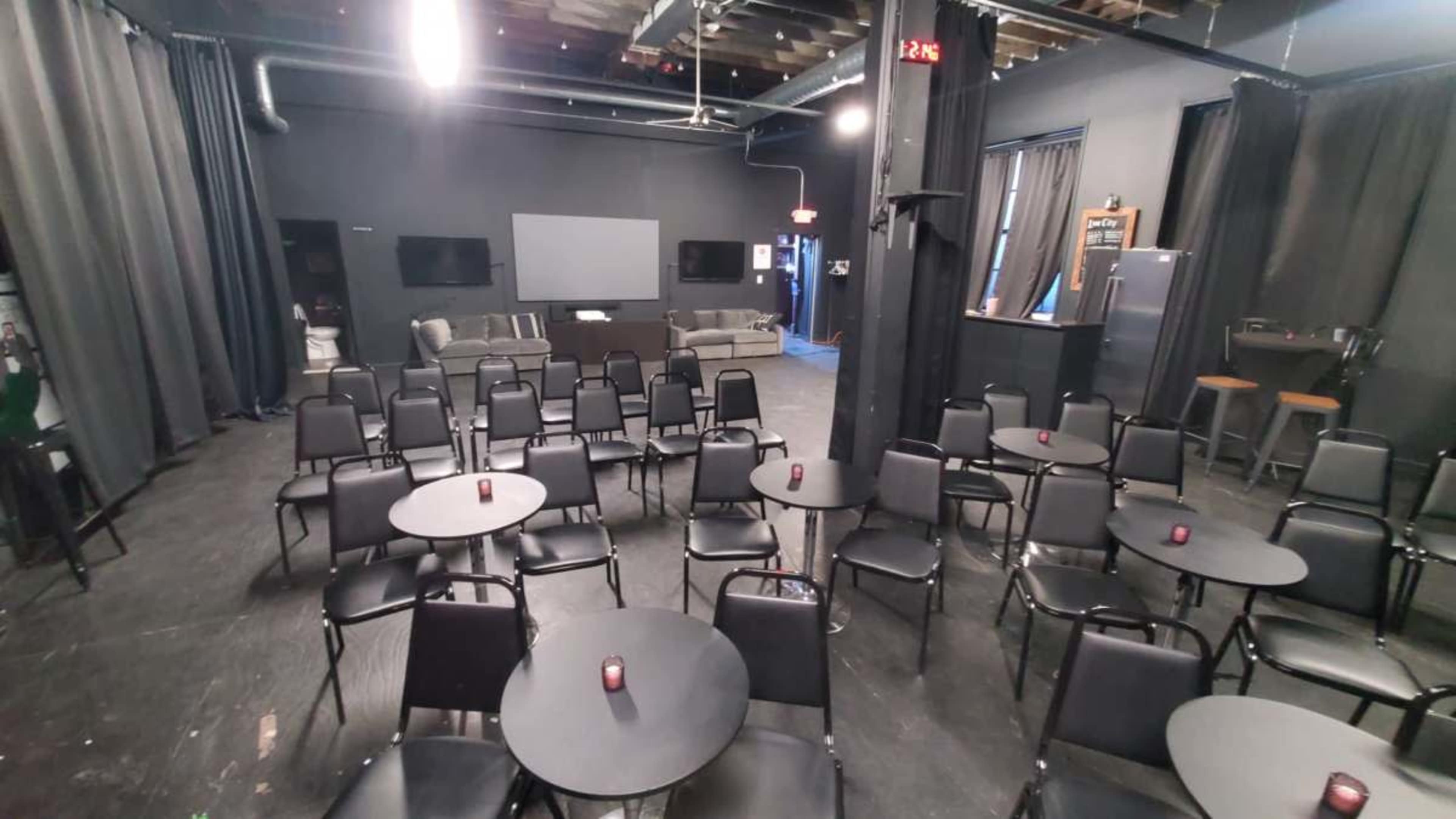 The image shows a spacious indoor venue set up with rows of black tables and chairs facing a screen, with a bar area and lounge seating in the background.