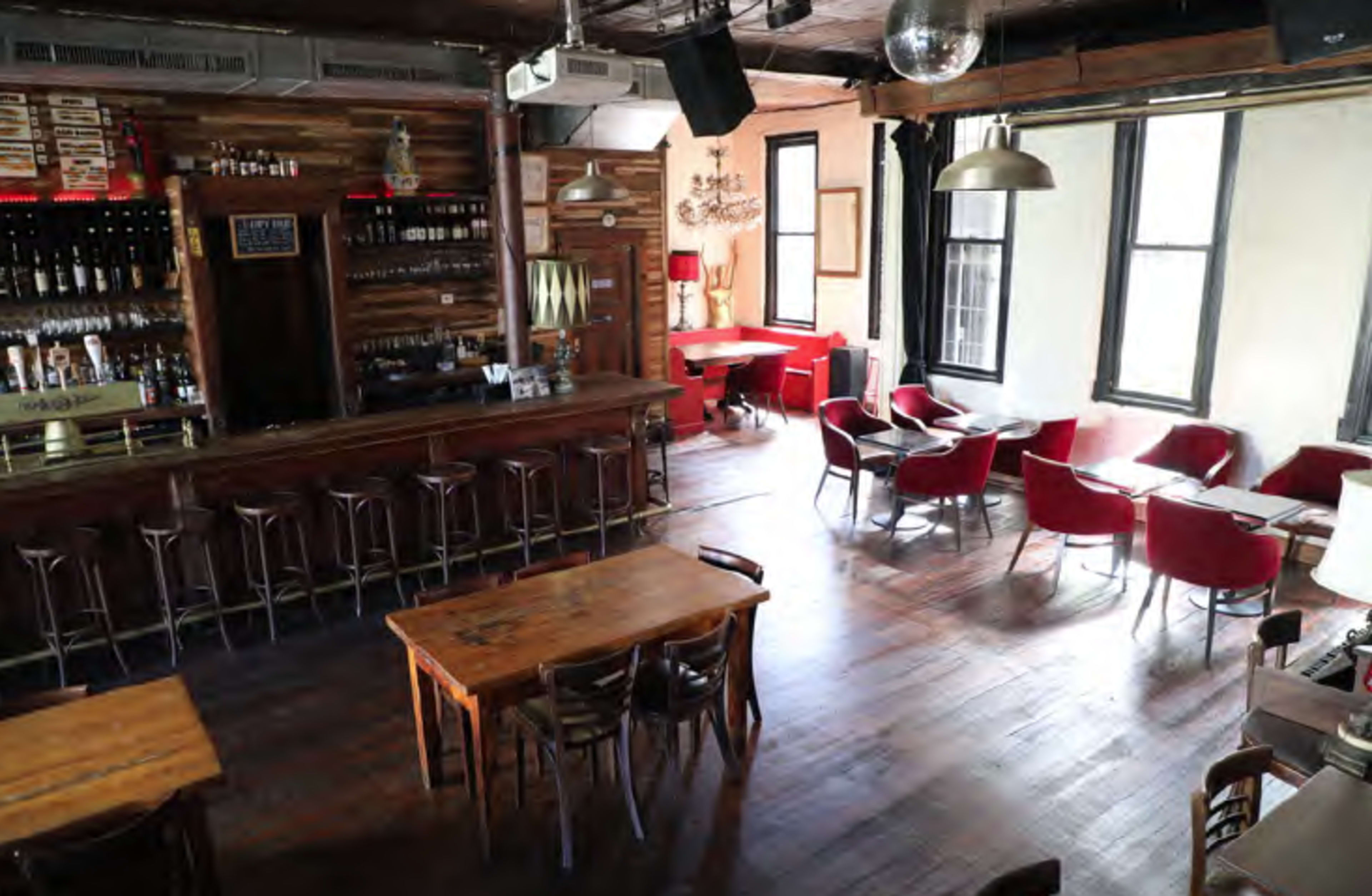 The image shows a spacious bar and dining area with wooden furniture, a long bar counter, and several red chairs arranged around tables.
