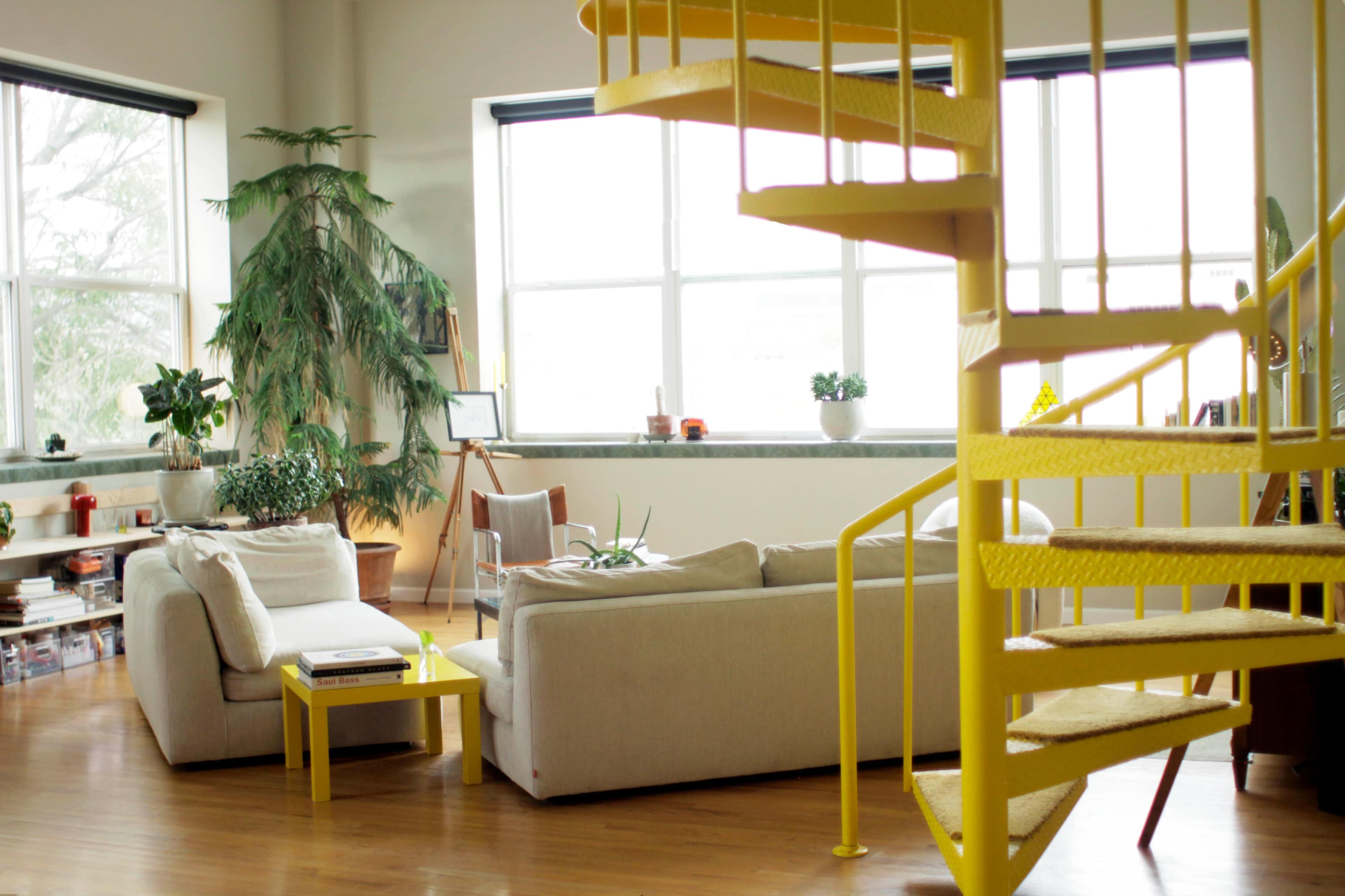 A bright living room features a white sofa, a yellow coffee table, and a spiral staircase, with large windows and indoor plants.
