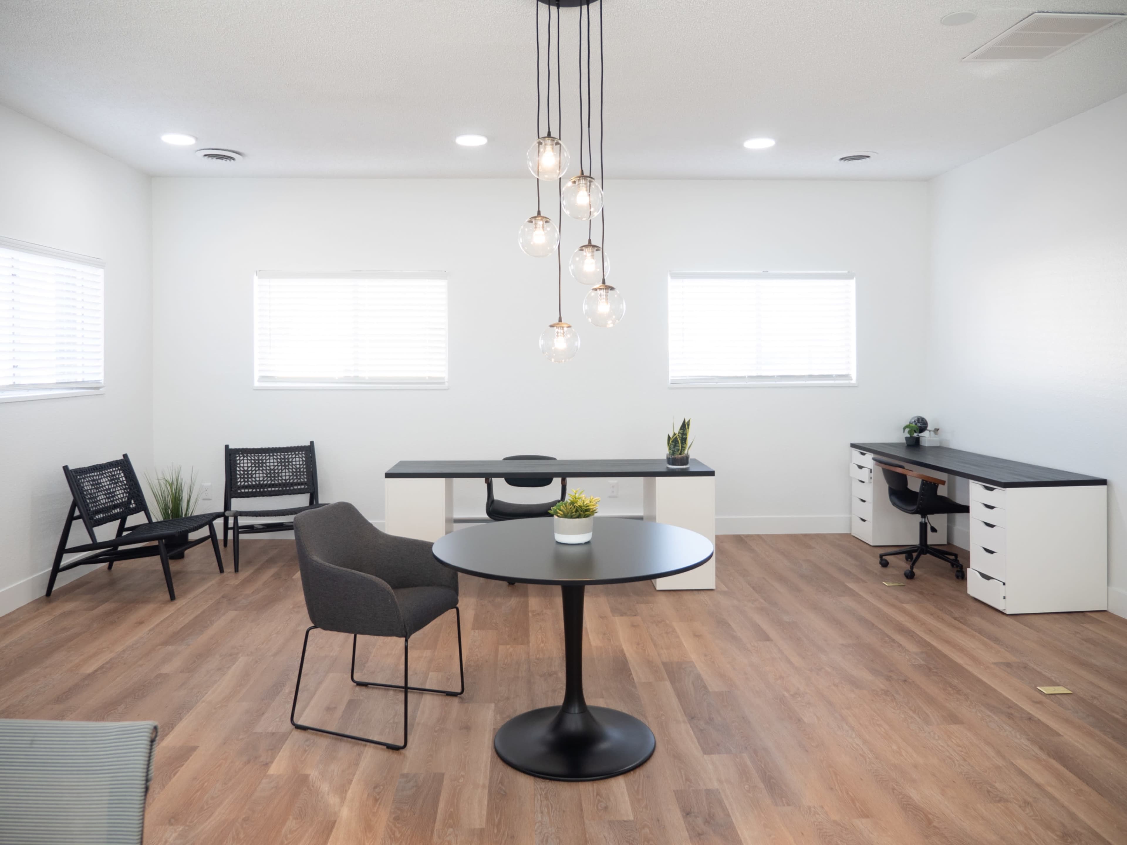 The image shows a modern office space featuring two desks, a round table, and black chairs, with large windows allowing natural light to enter.