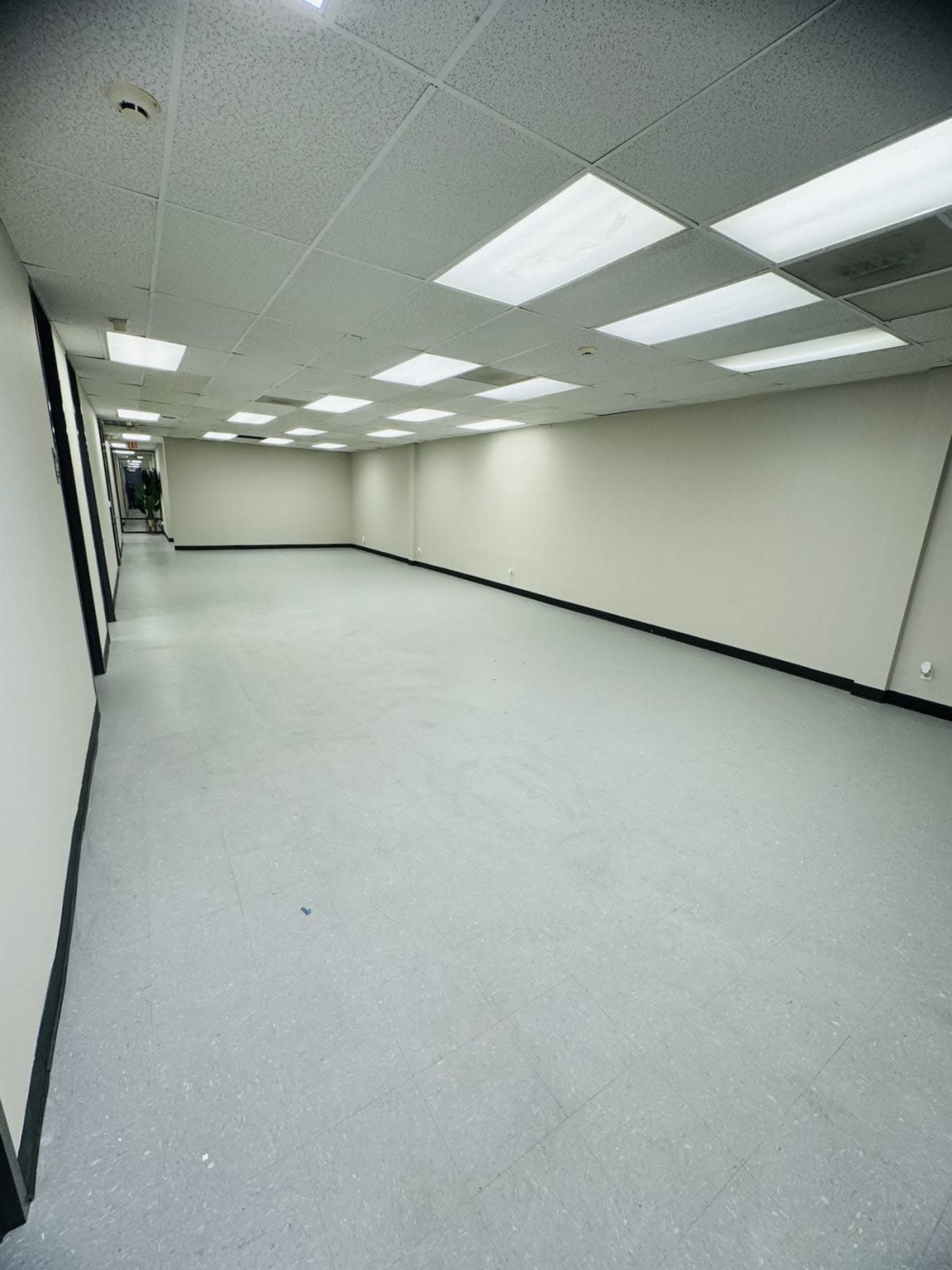 The image shows a large, empty hallway with white walls and fluorescent ceiling lights.