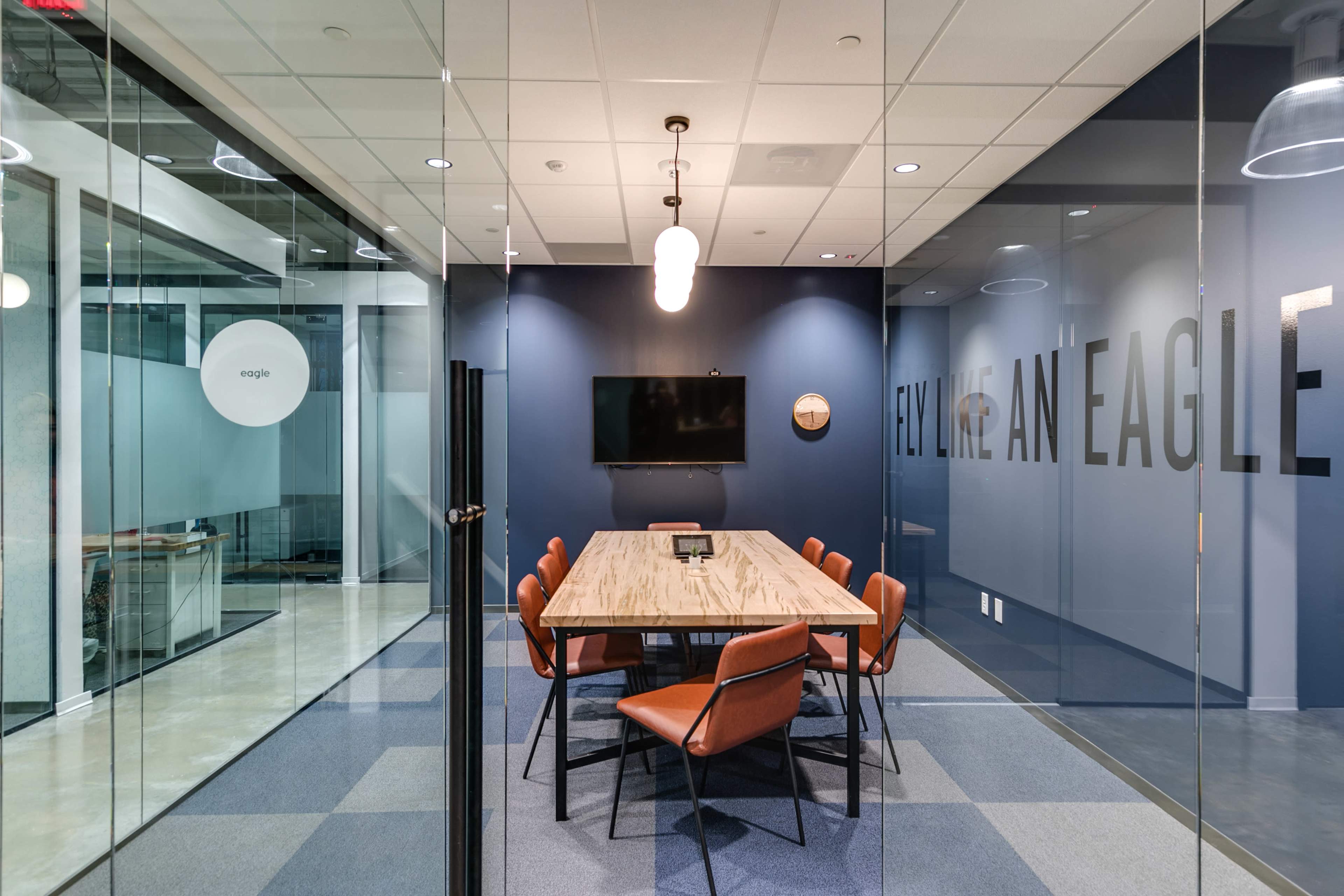 A conference room features a wooden table surrounded by orange chairs, with a wall displaying the phrase "FLY LIKE AN EAGLE" and a television mounted on the wall.