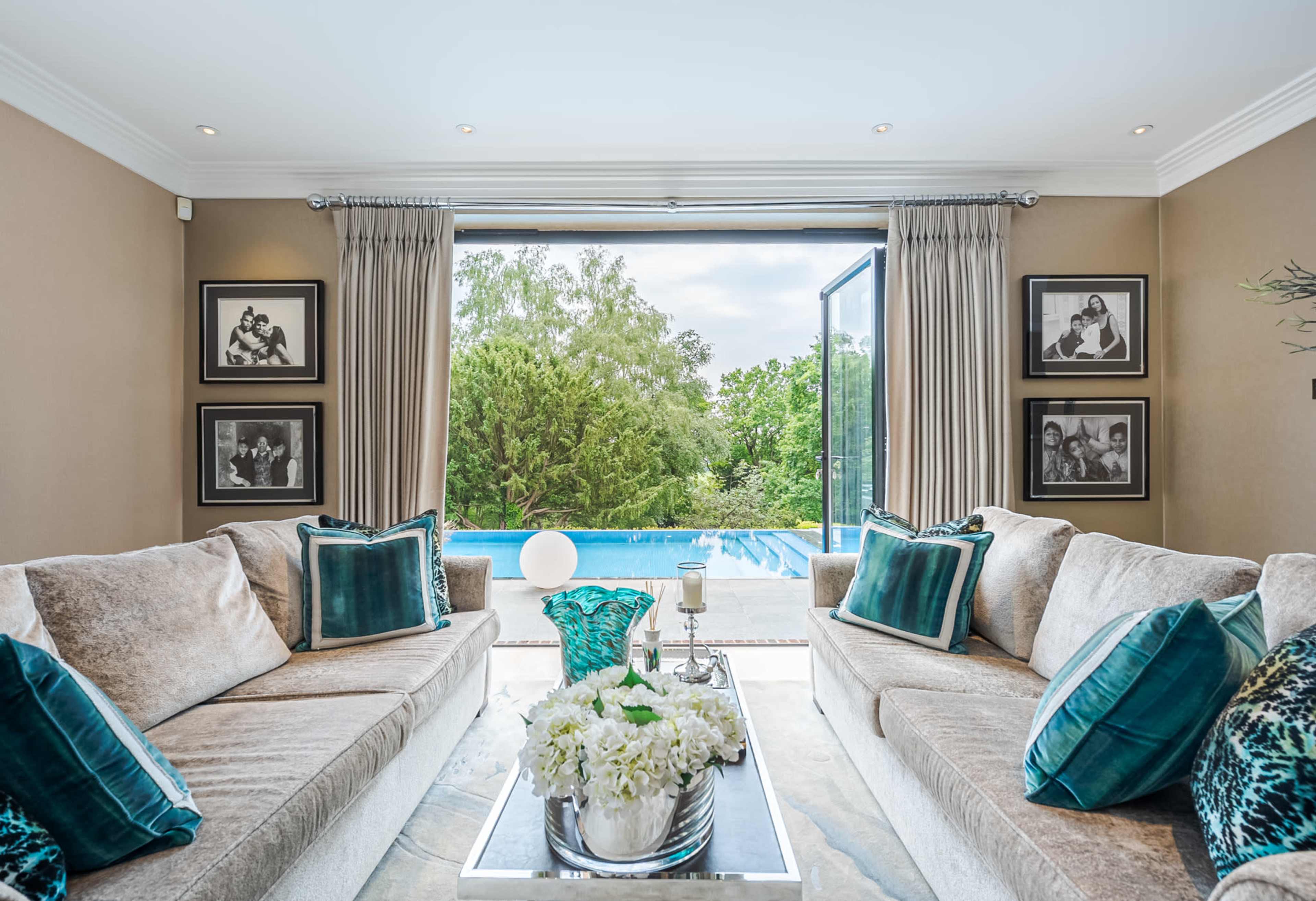 The living room features two sofas with teal accent pillows, a glass coffee table, and large windows opening to a poolside view.