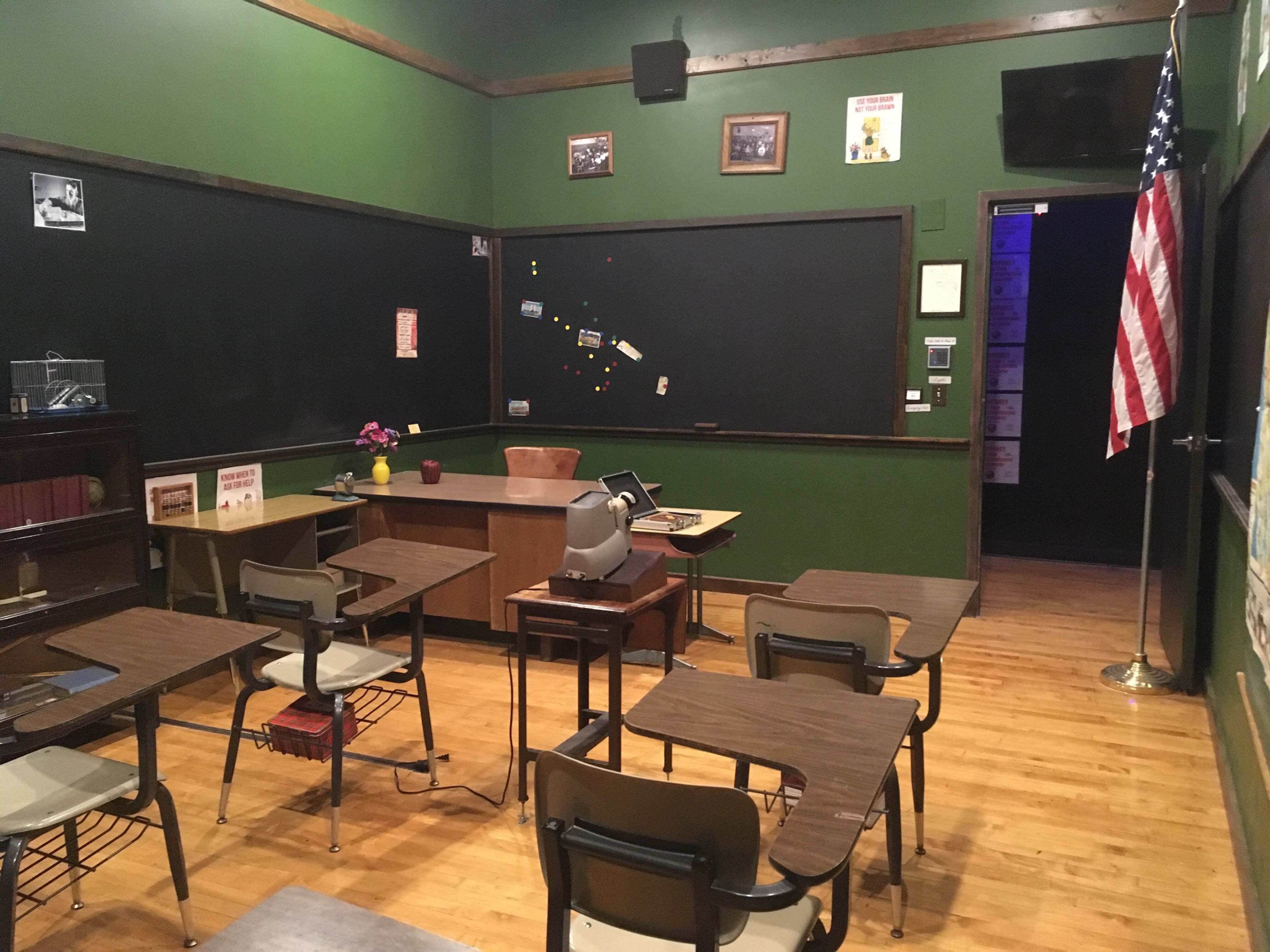 A vintage classroom features wooden desks, a chalkboard, and a desk at the front, with an American flag positioned in the corner.