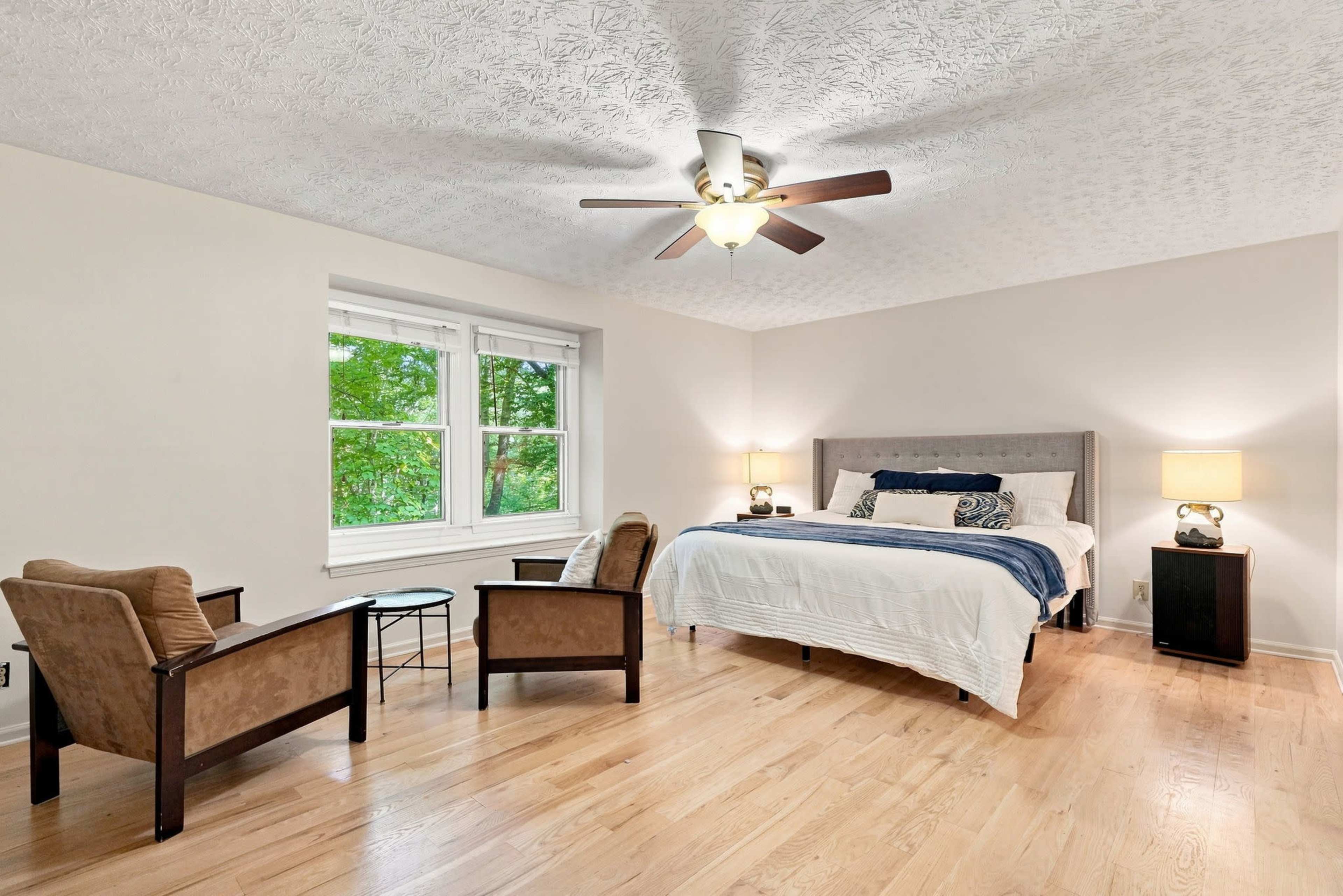 The image shows a well-lit bedroom featuring a bed with a gray headboard, two brown armchairs, a small glass table, and a ceiling fan.