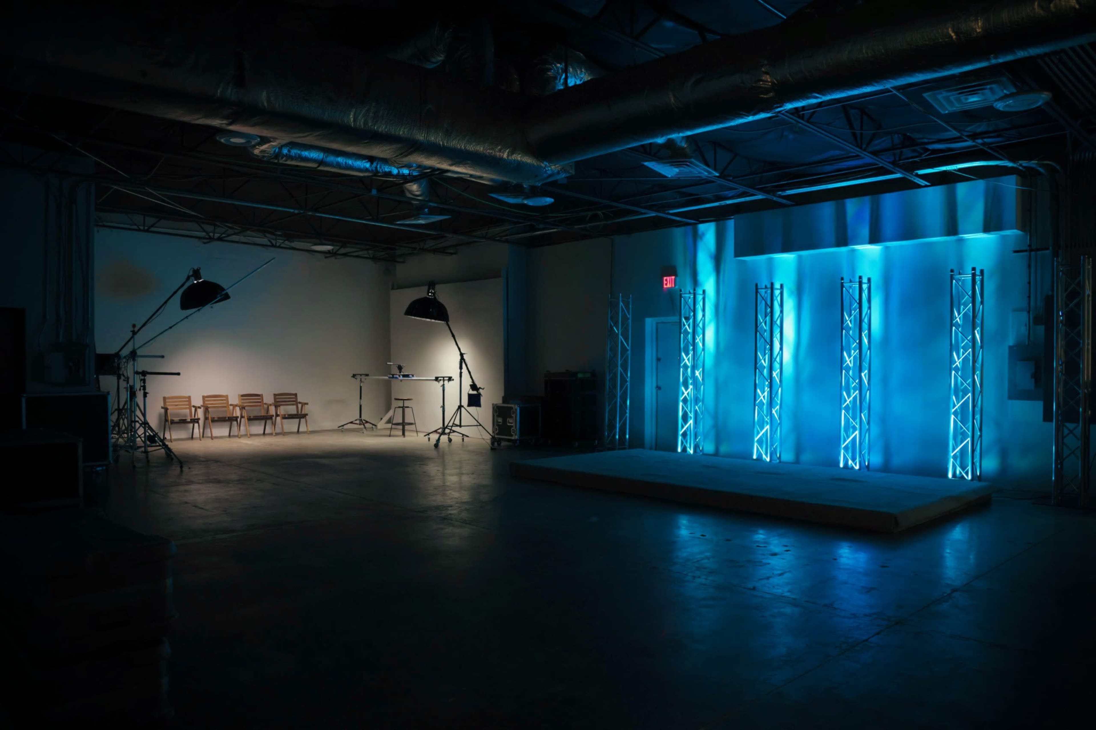 A dimly lit studio features a stage with blue lighting, flanked by four vertical truss towers, and a row of empty chairs facing a table with equipment.
