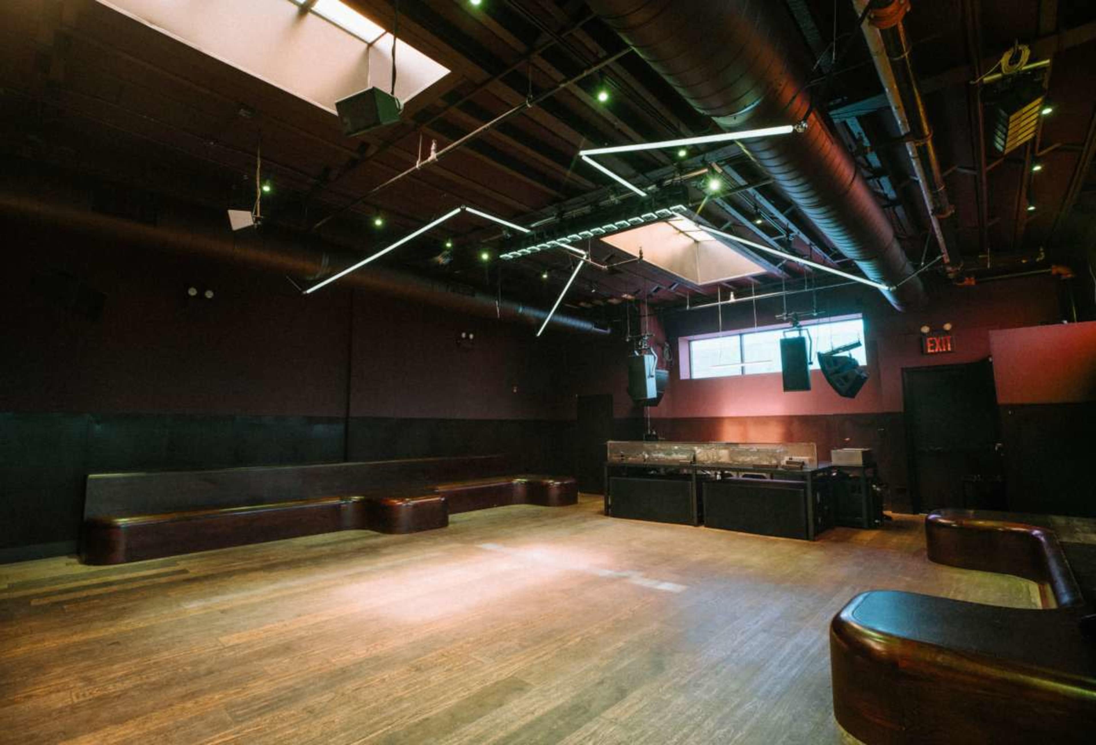 The image shows an empty event space featuring wooden benches, dark walls, and modern lighting fixtures.