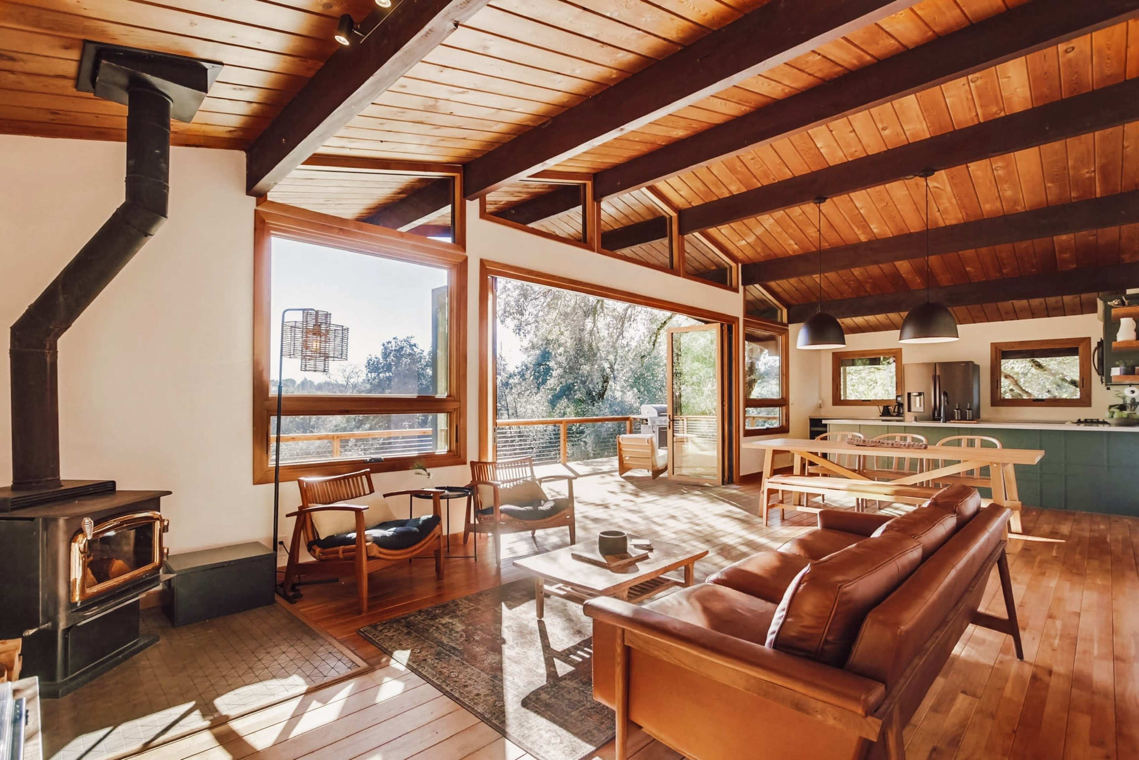 A modern, open-concept living space with a wood-burning stove, large windows, and a dining area leading to a deck.