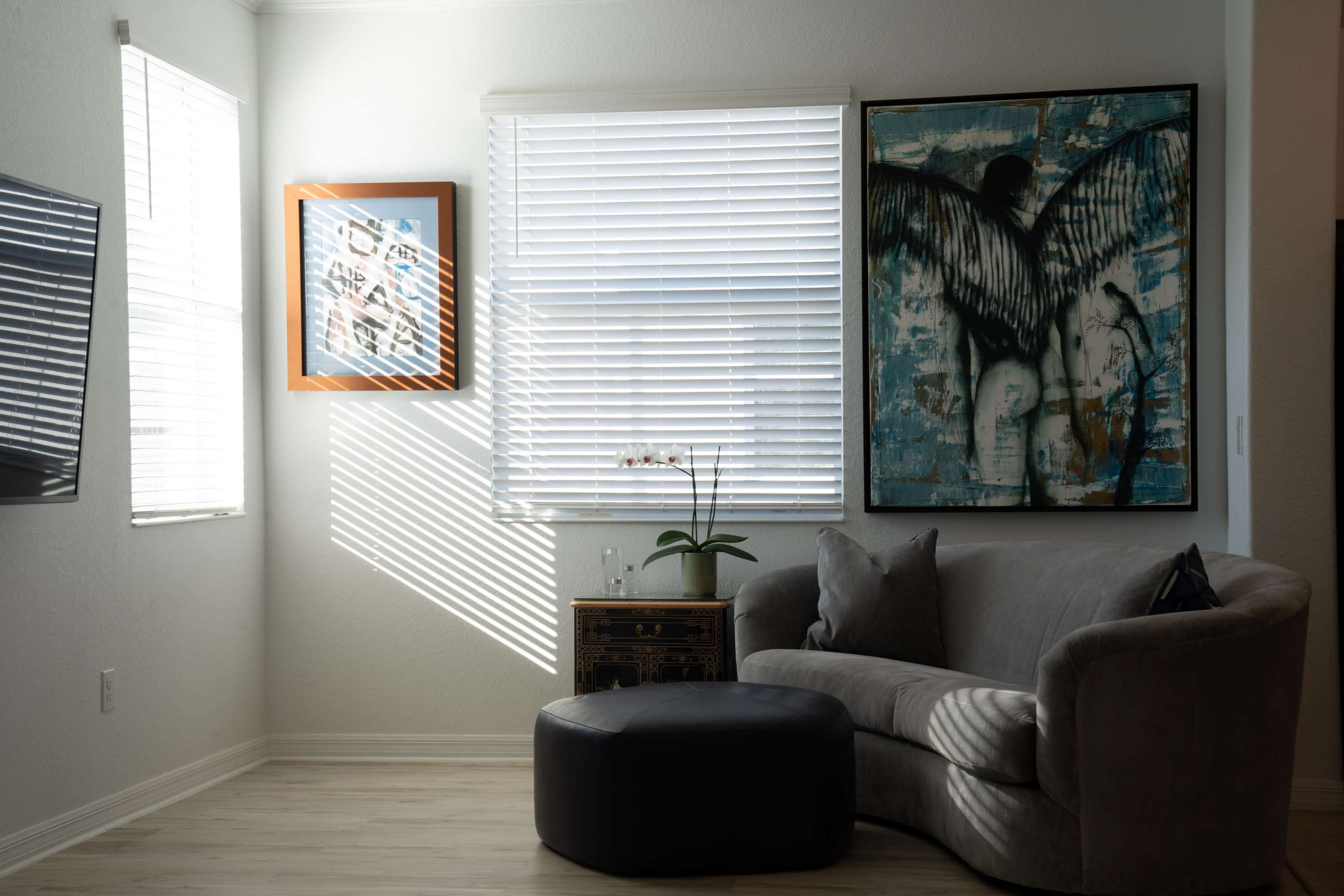 A bright living room corner featuring a gray couch, an ottoman, a decorative plant, and two framed artworks on the walls.