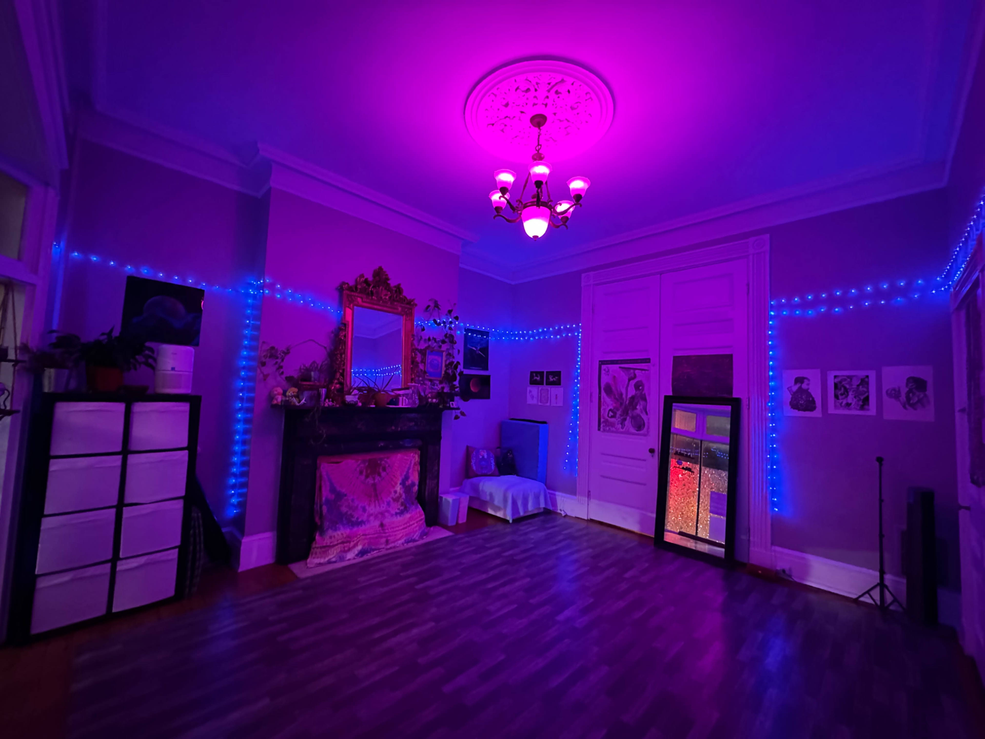 A dimly lit room features purple LED lights outlining the walls, a decorative fireplace, a large mirror, and various artworks.