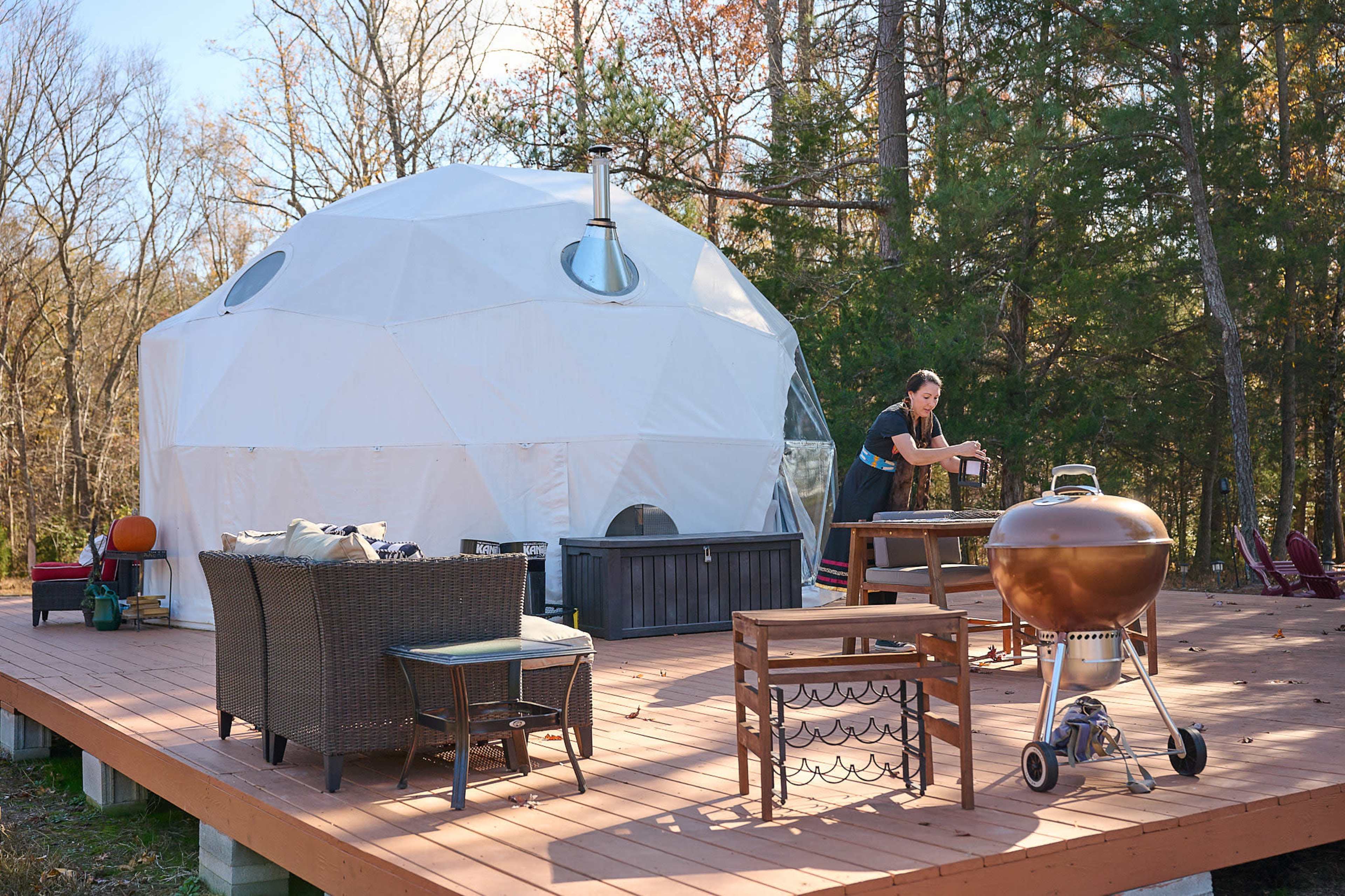 A person is preparing food at a table next to a geodesic dome structure on a wooden deck surrounded by trees.
