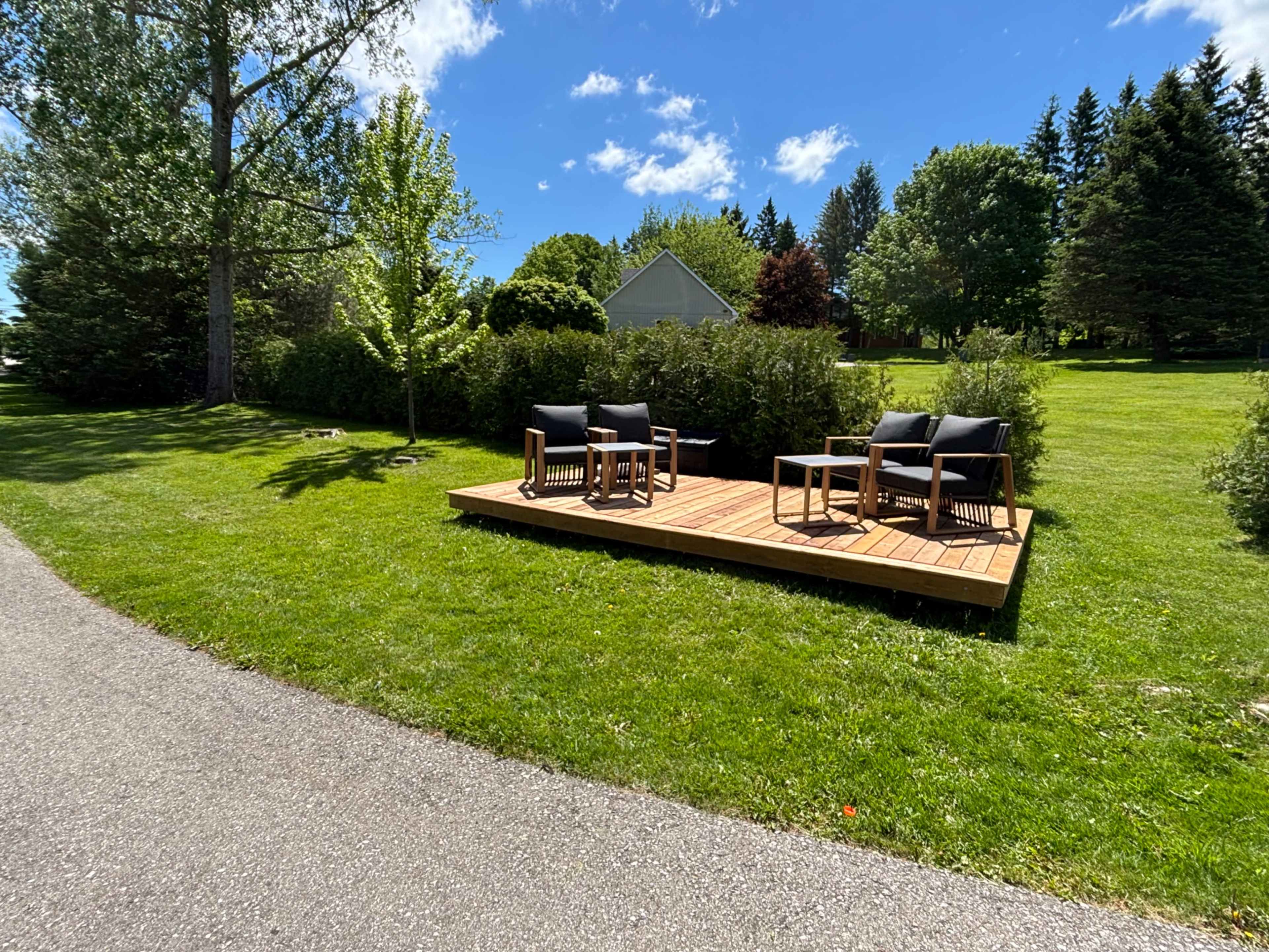 A wooden deck with two chairs is situated on a grassy area alongside a paved path, surrounded by trees and shrubs.