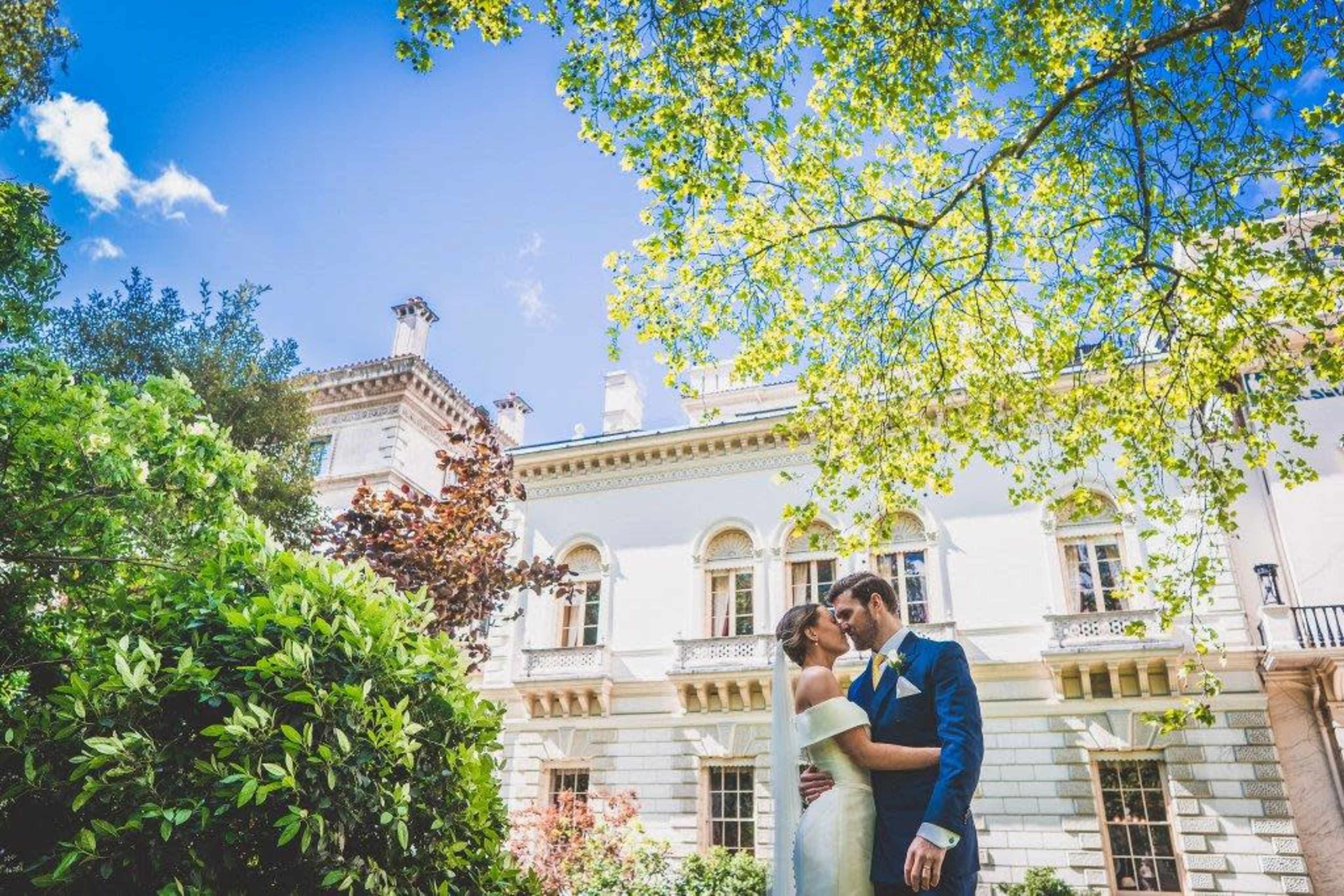 A bride and groom kiss in a lush garden in front of an elegant mansion.