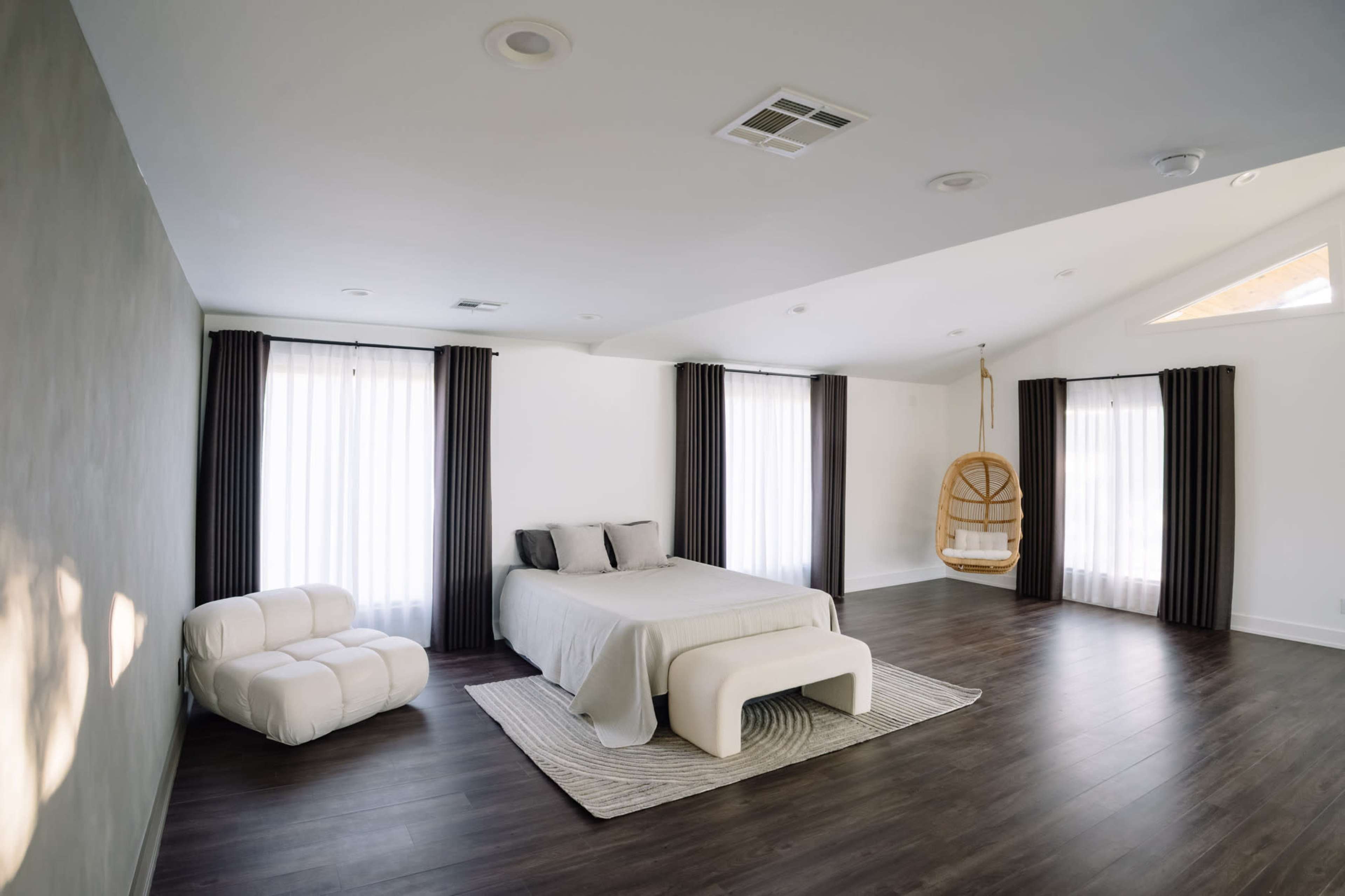 The image shows a spacious, minimalist bedroom featuring a bed, a white lounge chair, and a hanging chair, with large windows dressed in sheer curtains.