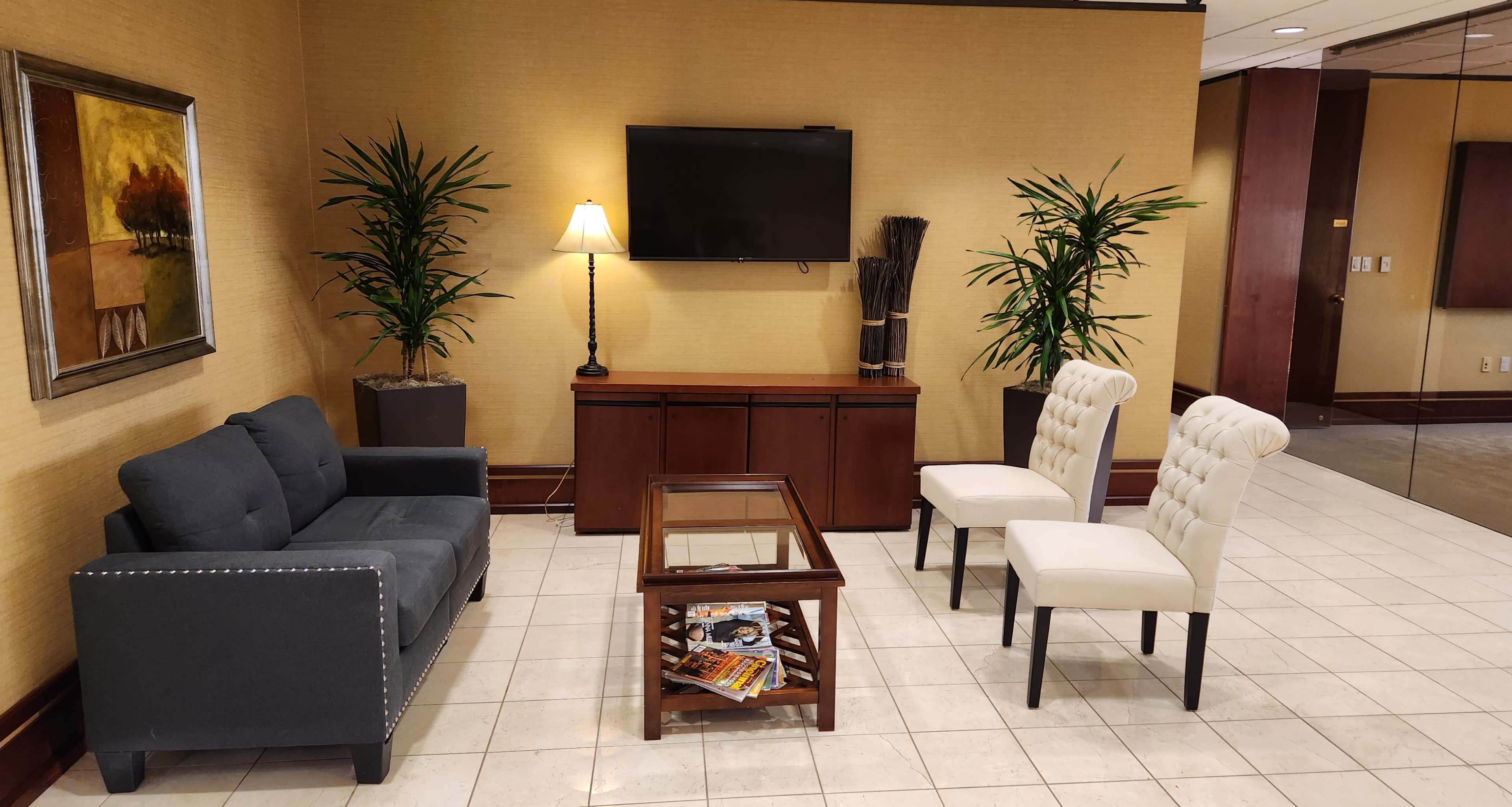 A waiting area features a gray couch, two white chairs, a wooden coffee table with magazines, a lamp, and potted plants against a beige wall with a painting.
