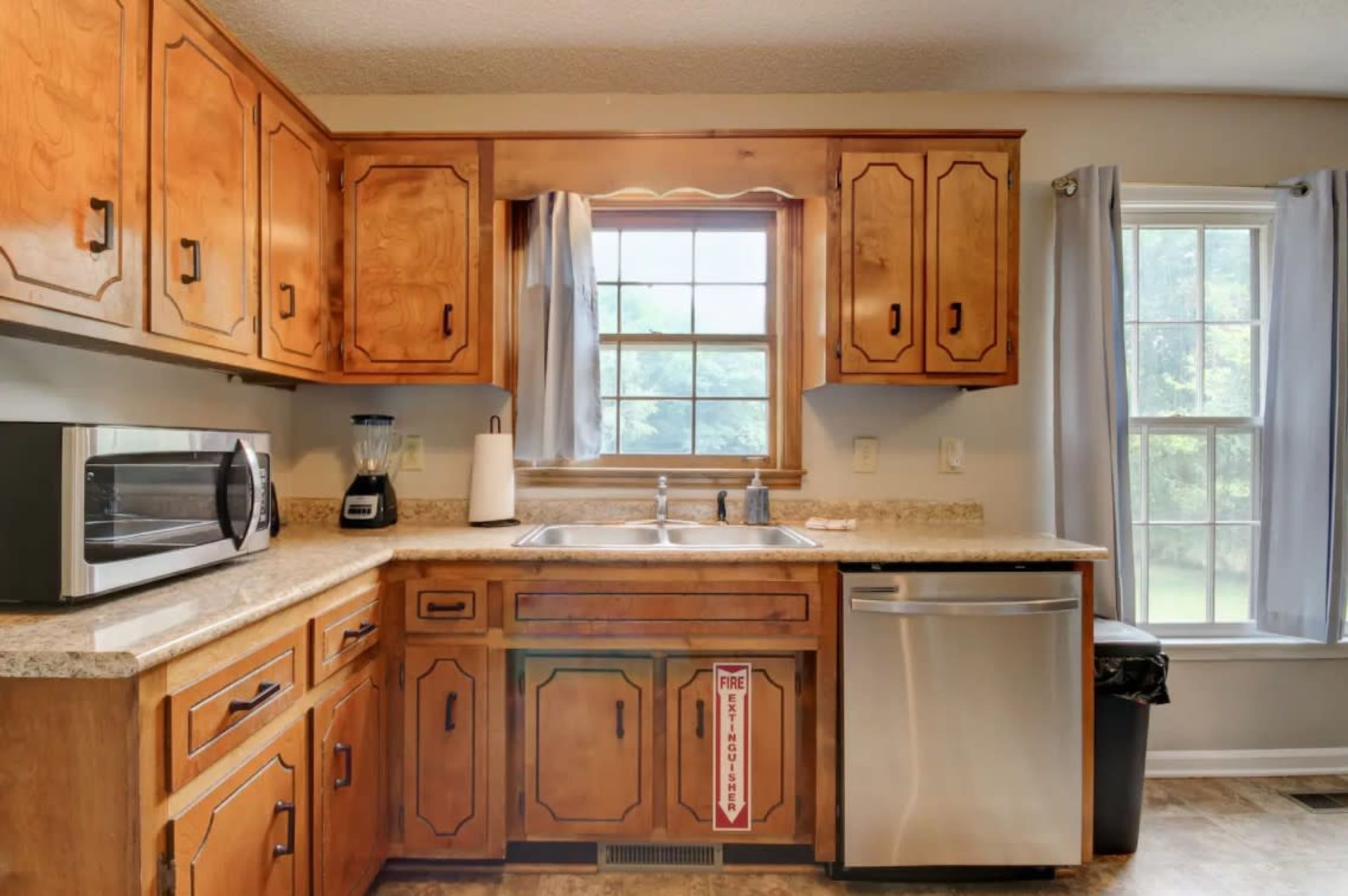 The image shows a kitchen with wooden cabinets, a granite countertop, a sink under a window, a microwave, a blender, a dishwasher, and a trash bin.