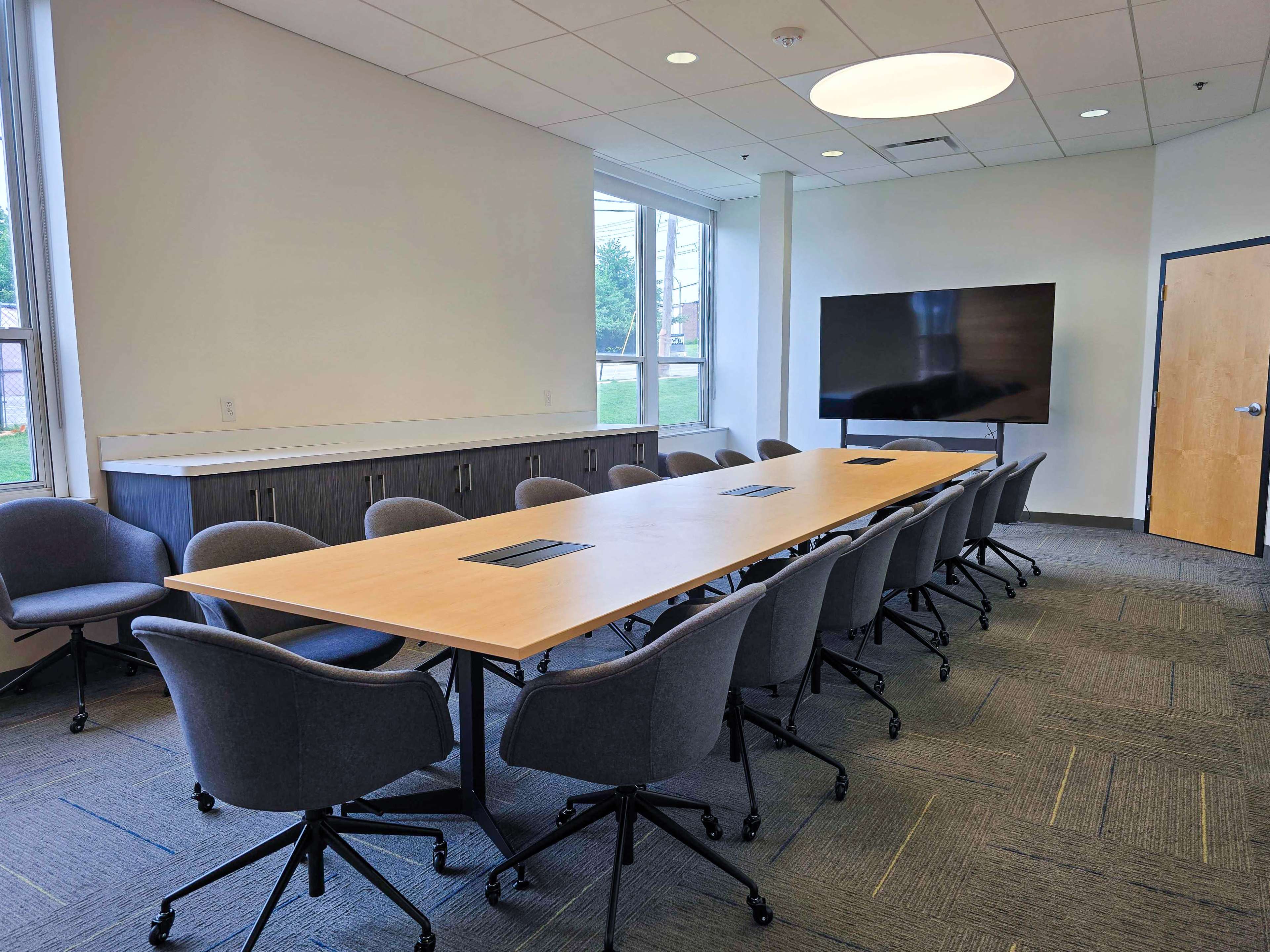 The image shows a modern conference room with a long table surrounded by rolling chairs and a large screen mounted on the wall.