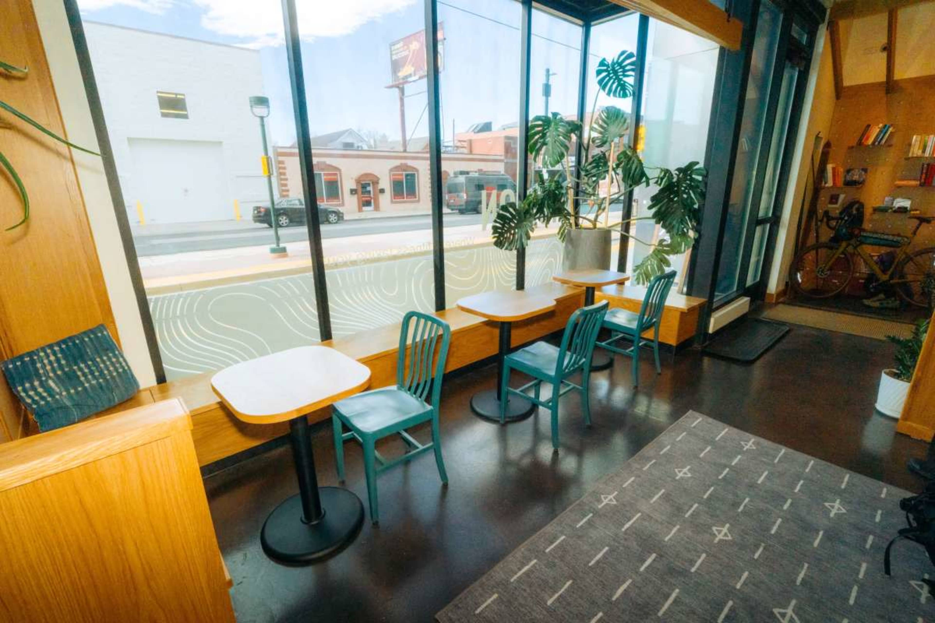The image shows a modern café area with small tables and colorful chairs, large windows allowing natural light, and potted plants in the background.