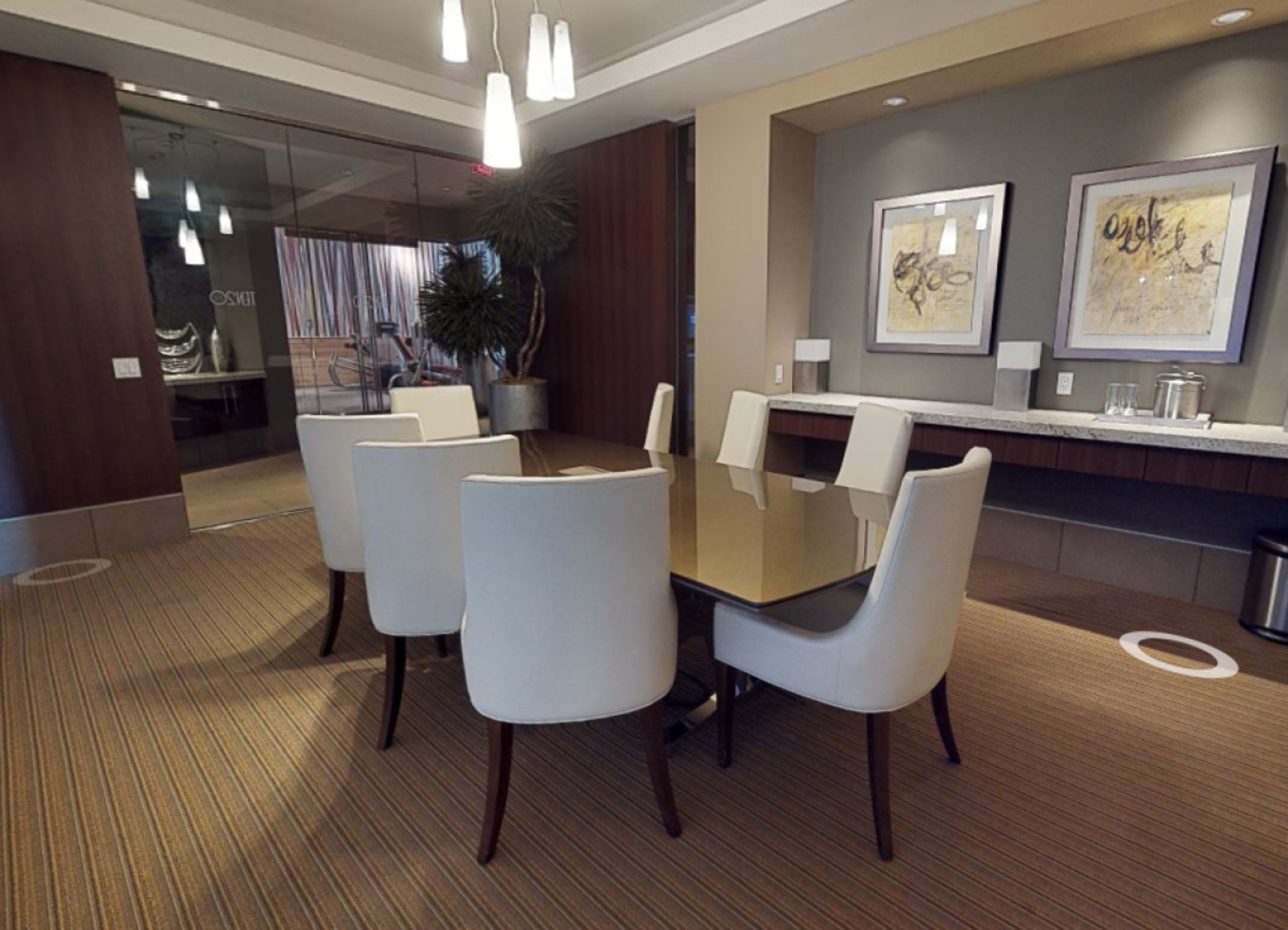 A conference room features a rectangular table surrounded by white chairs, with a countertop and framed artwork on the walls.